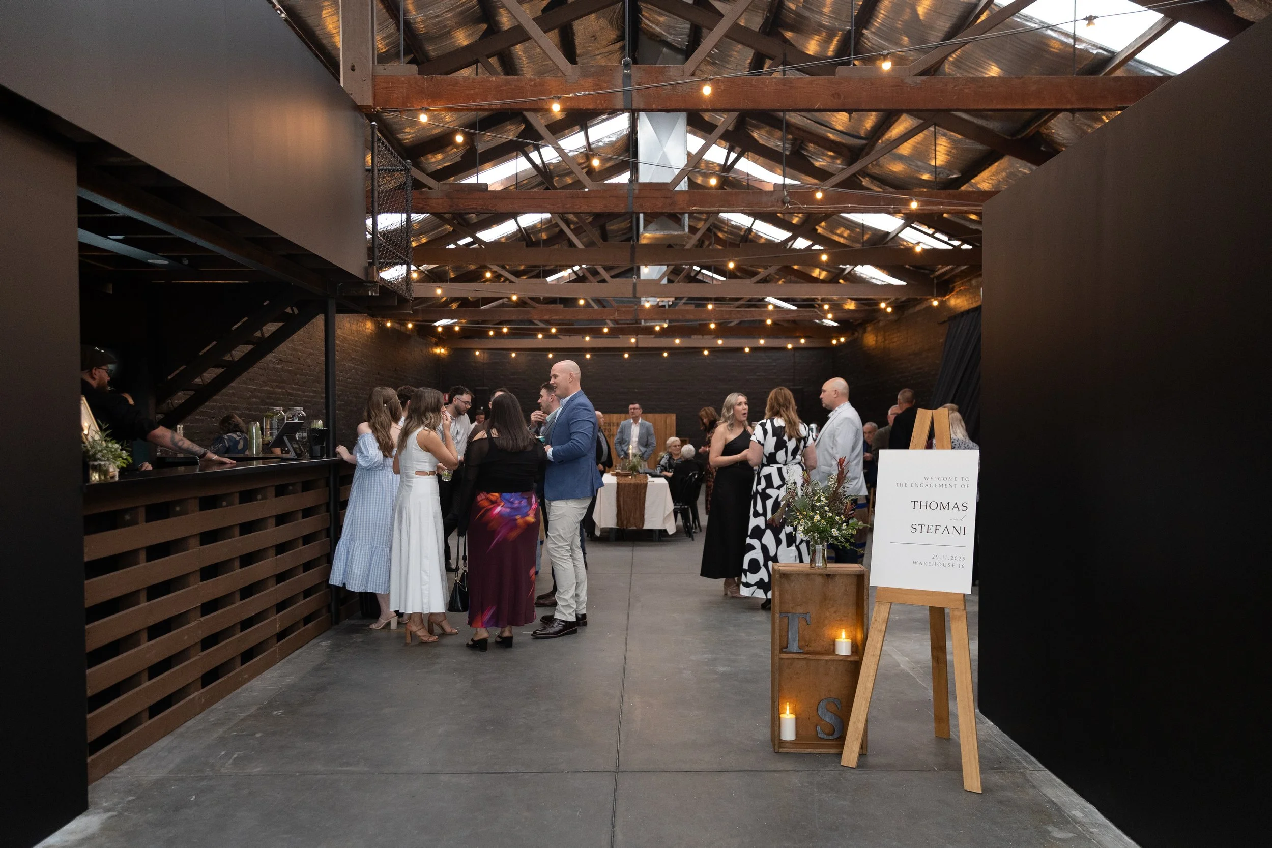 Group of people at a wedding reception inside a rustic-industrial venue with string lights, wooden beams, and a sign indicating it is for Thomas and Stefani's celebration on November 29, 2025.
