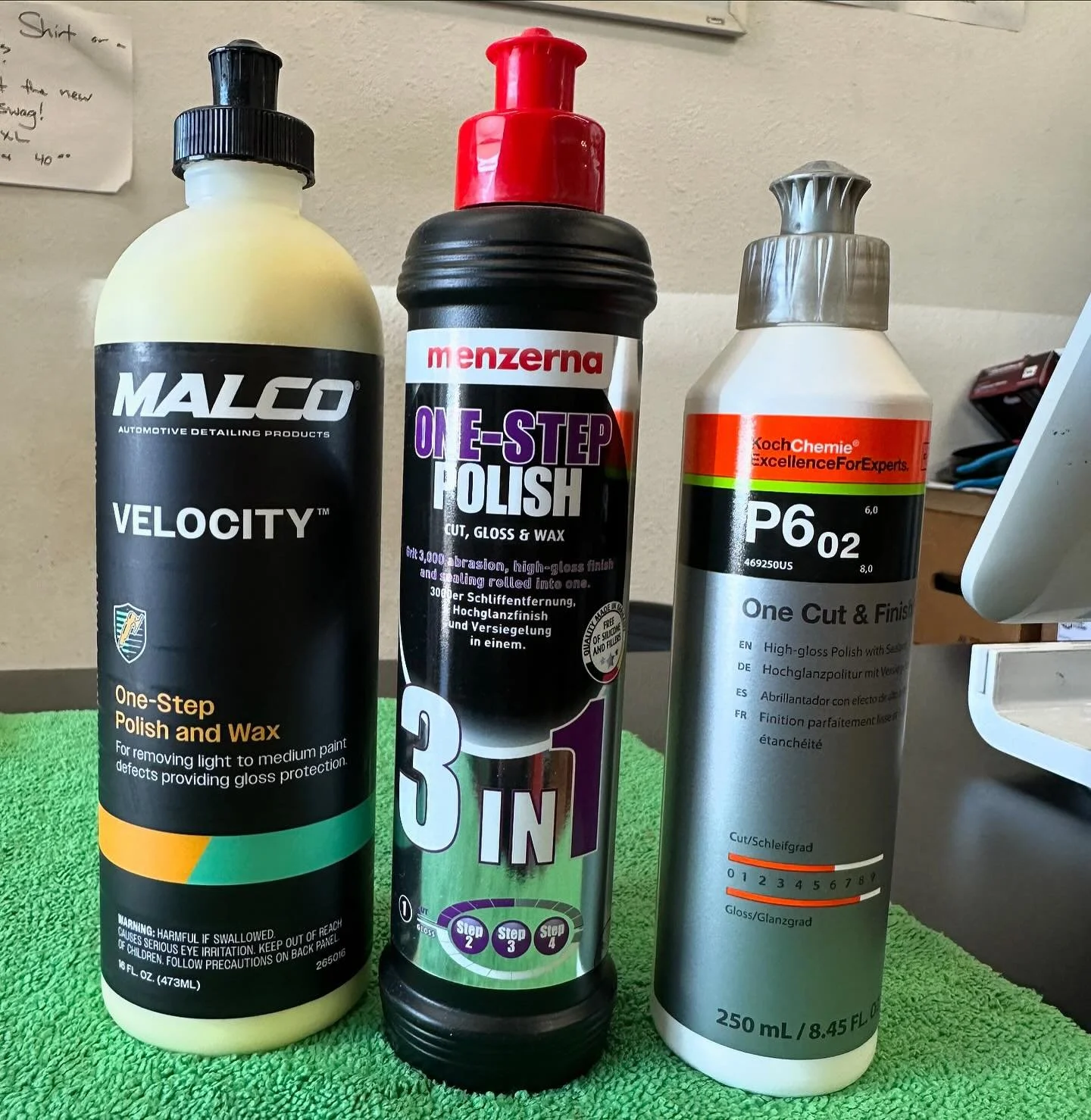 Three bottles of automotive detailing products on a green textured surface, against a beige wall. The left bottle is black and yellow labeled Malco Velocity, a one-step polish and wax. The middle bottle is black with a red cap labeled Menzerna One E-Step Polish. The right bottle is silver with a gray cap labeled KochChemie P6 02, a high-gloss polish.