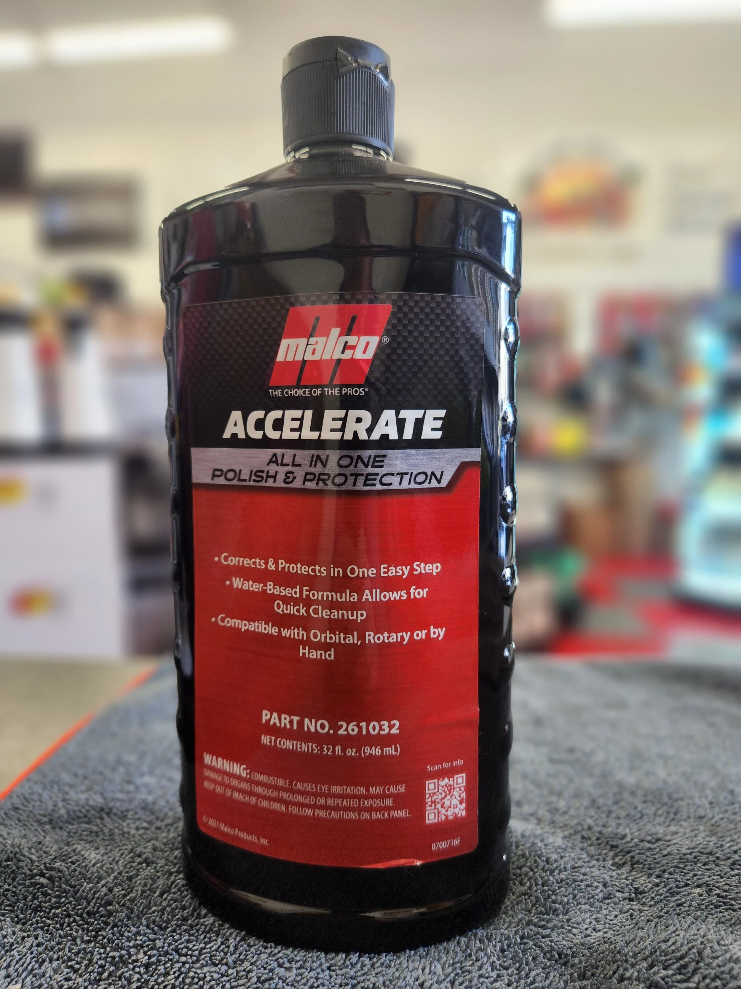 A black bottle of Malco Accelerate All-In-One Polish & Protection on a gray textured surface, with shelves and products blurry in the background.