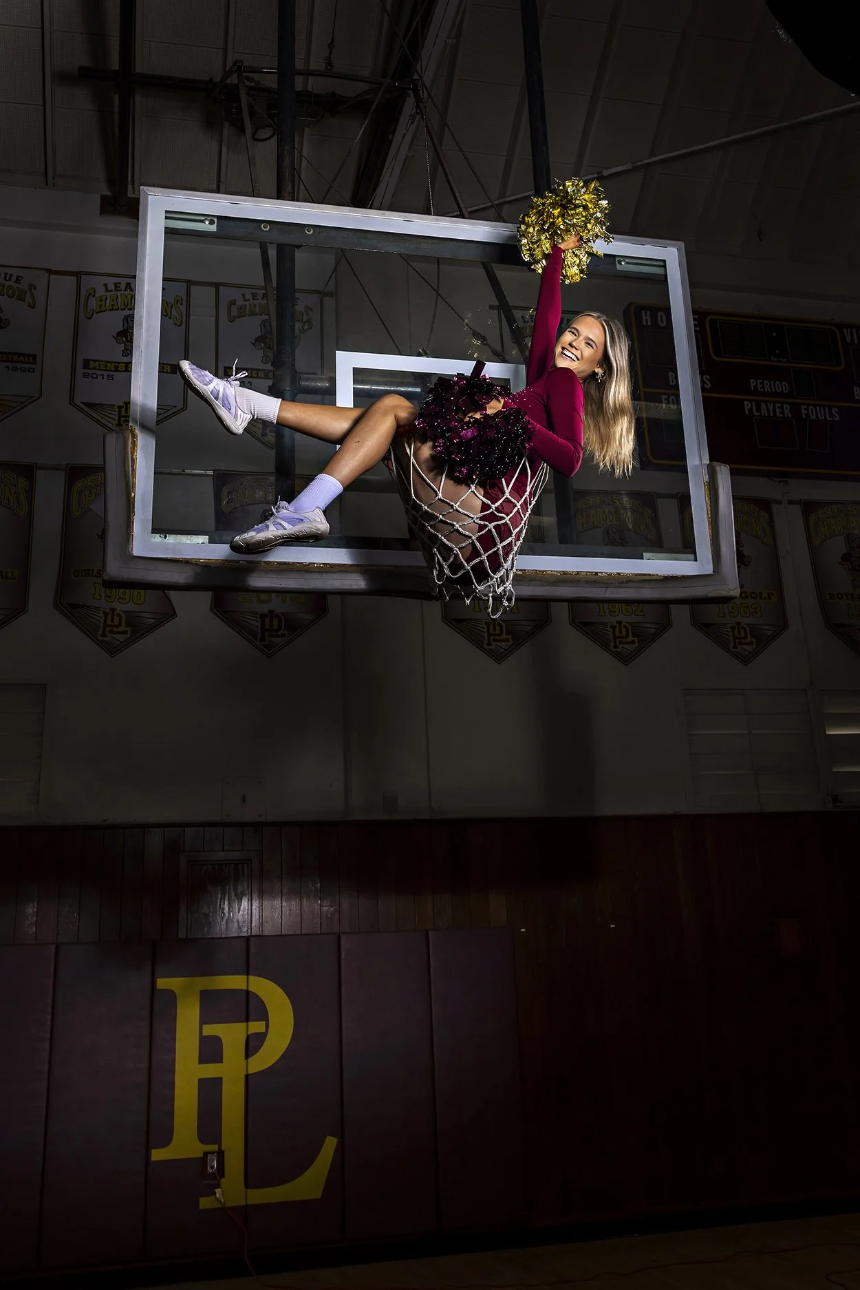 A woman dressed as a cheerleader sitting on a basketball hoop, holding gold pom-poms, inside a gymnasium with banners on the wall.