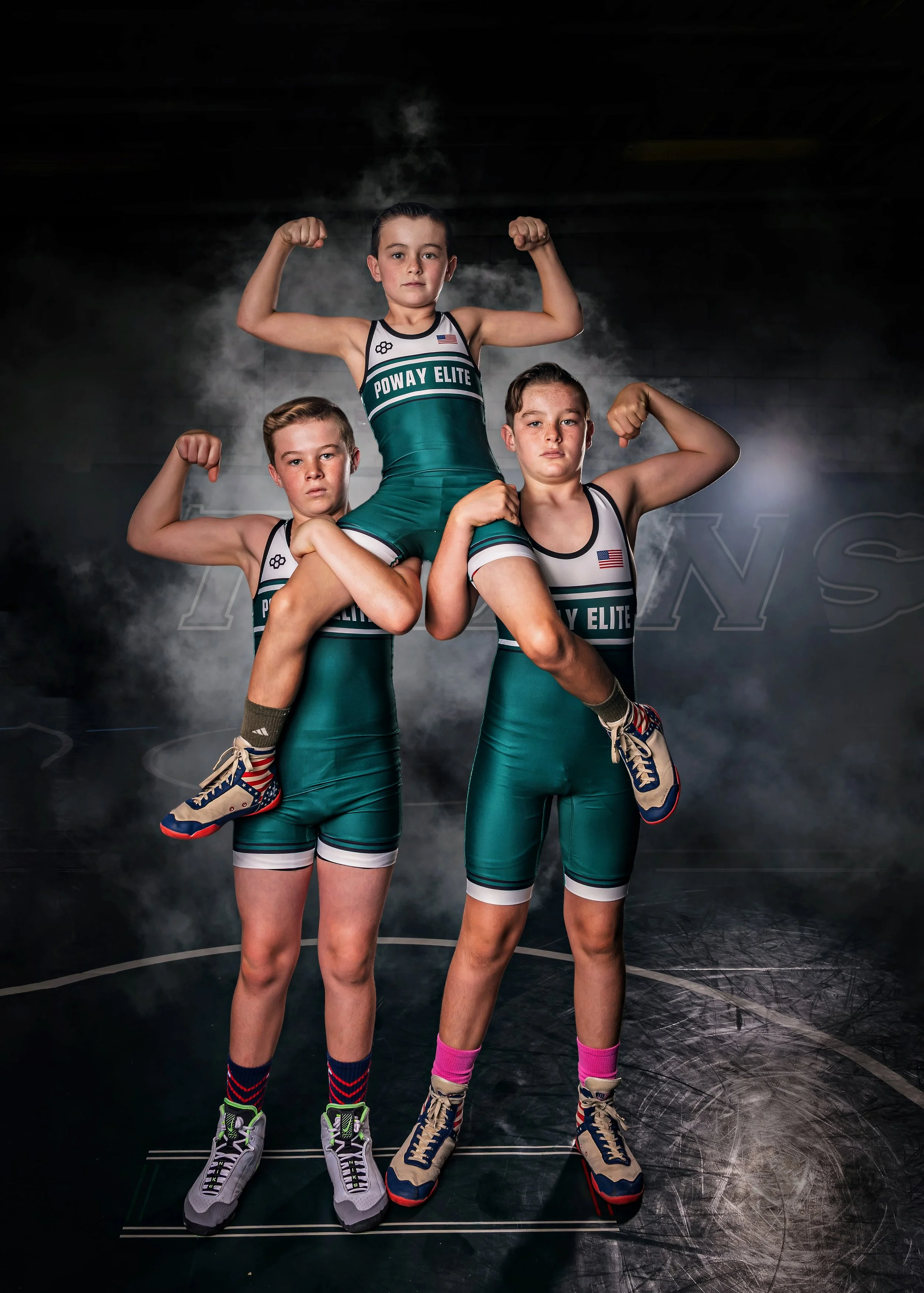 Three young boys in wrestling singlets posing with their arms flexed. The boy in the middle is lifted onto the shoulders of the other two boys. The background is dark with a smoky effect, and the boys are standing on a wrestling mat.