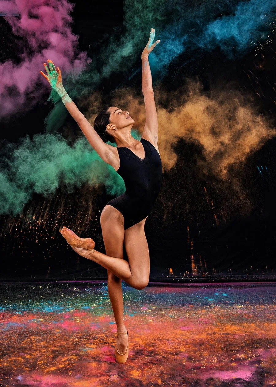 A ballet dancer performing a pose on stage with colorful powder and smoke in the background.
