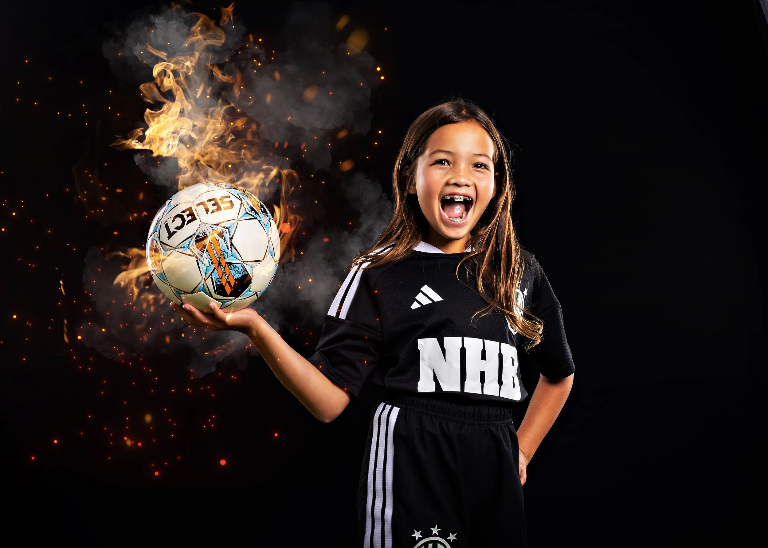 Young girl wearing a sports jersey holding a soccer ball on fire against a black background.