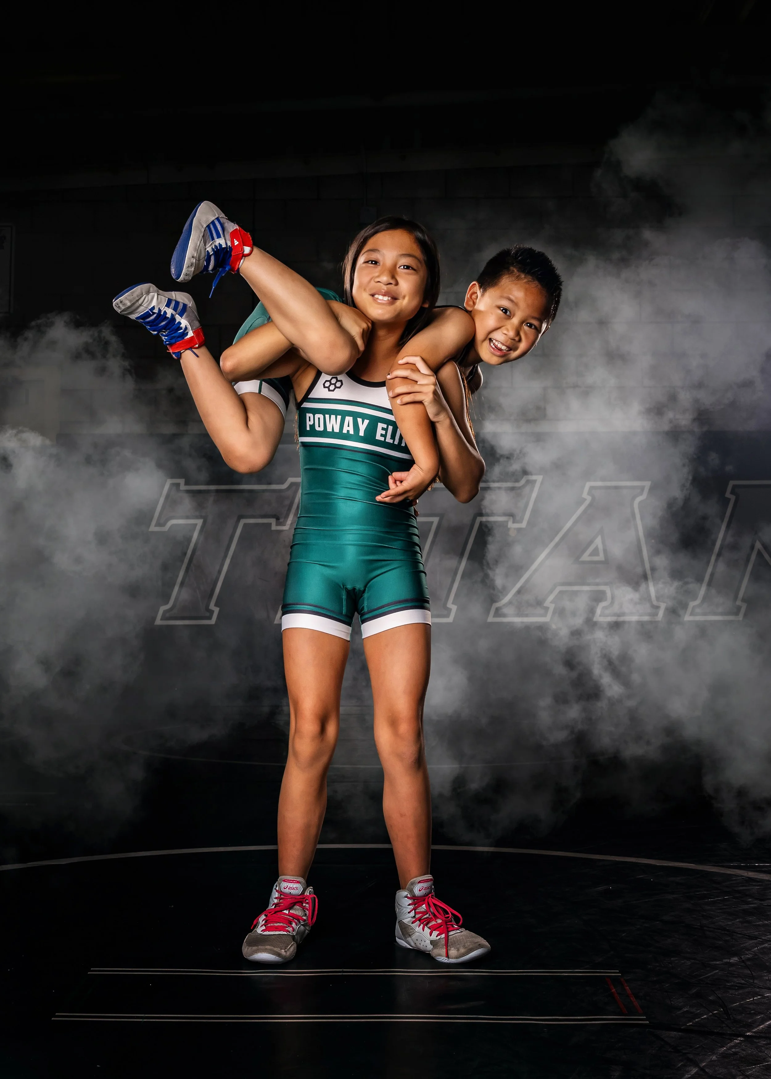 Young boy and girl sports enthusiasts, with girl in a track uniform carrying boy on her back in a gym or sports studio with a misty or smoky background.