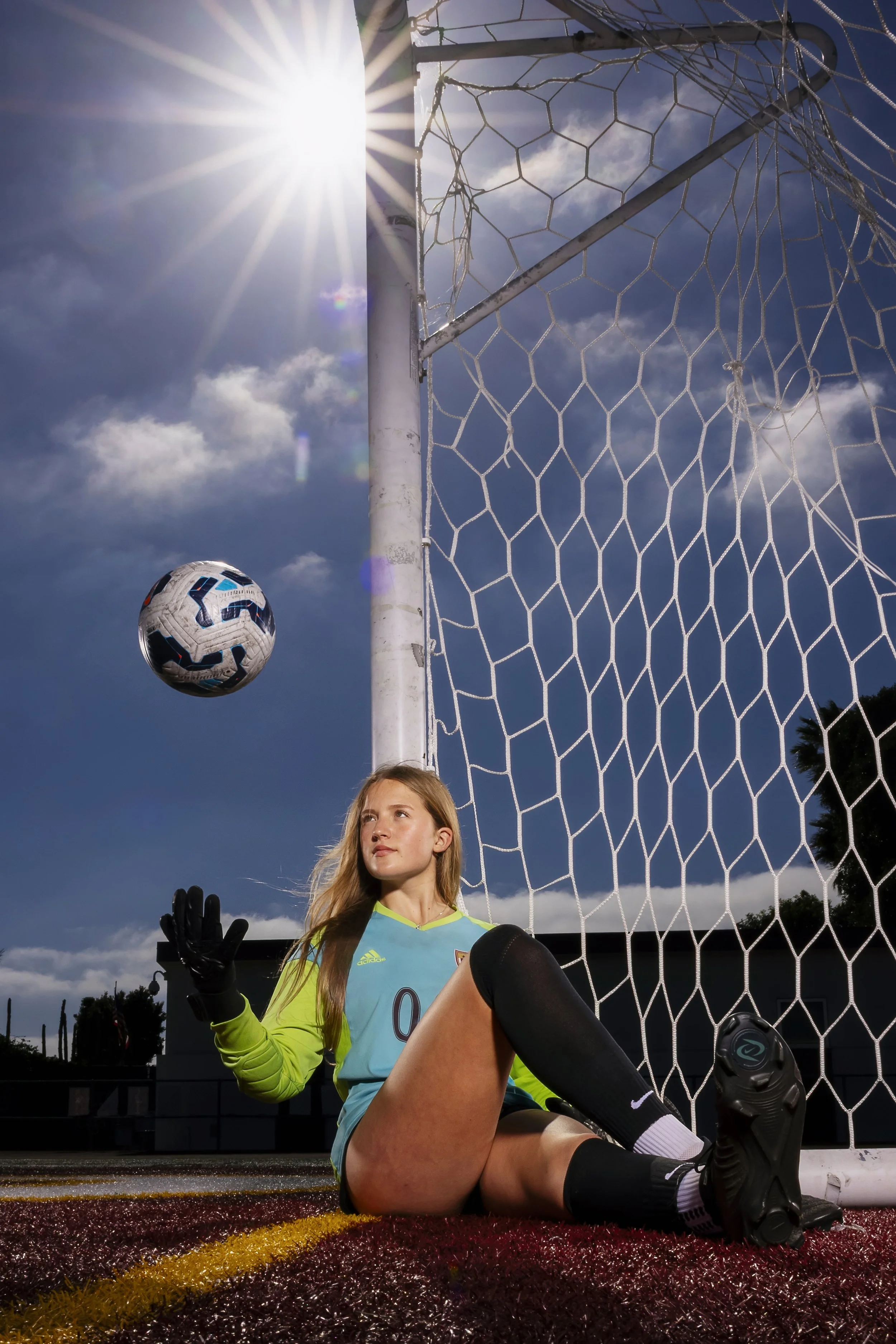 A female soccer goalie sitting on the ground, wearing a light blue jersey, black gloves, and black knee-high socks, with a soccer ball in the air near the goalpost under the bright sun and a partly cloudy sky.