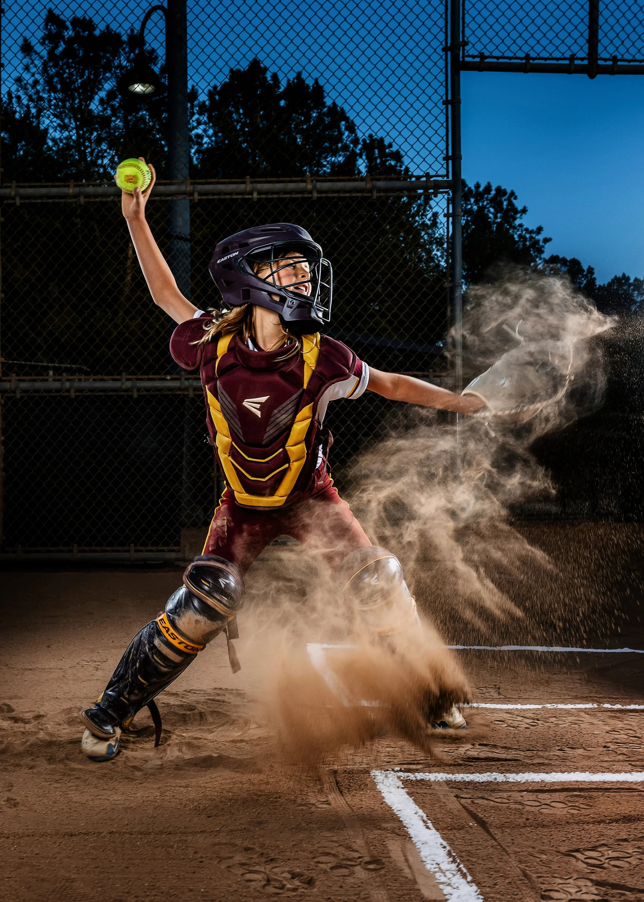 A female baseball player in full gear, including helmet and catcher's armor, throwing a baseball from a dirt field during evening hours.