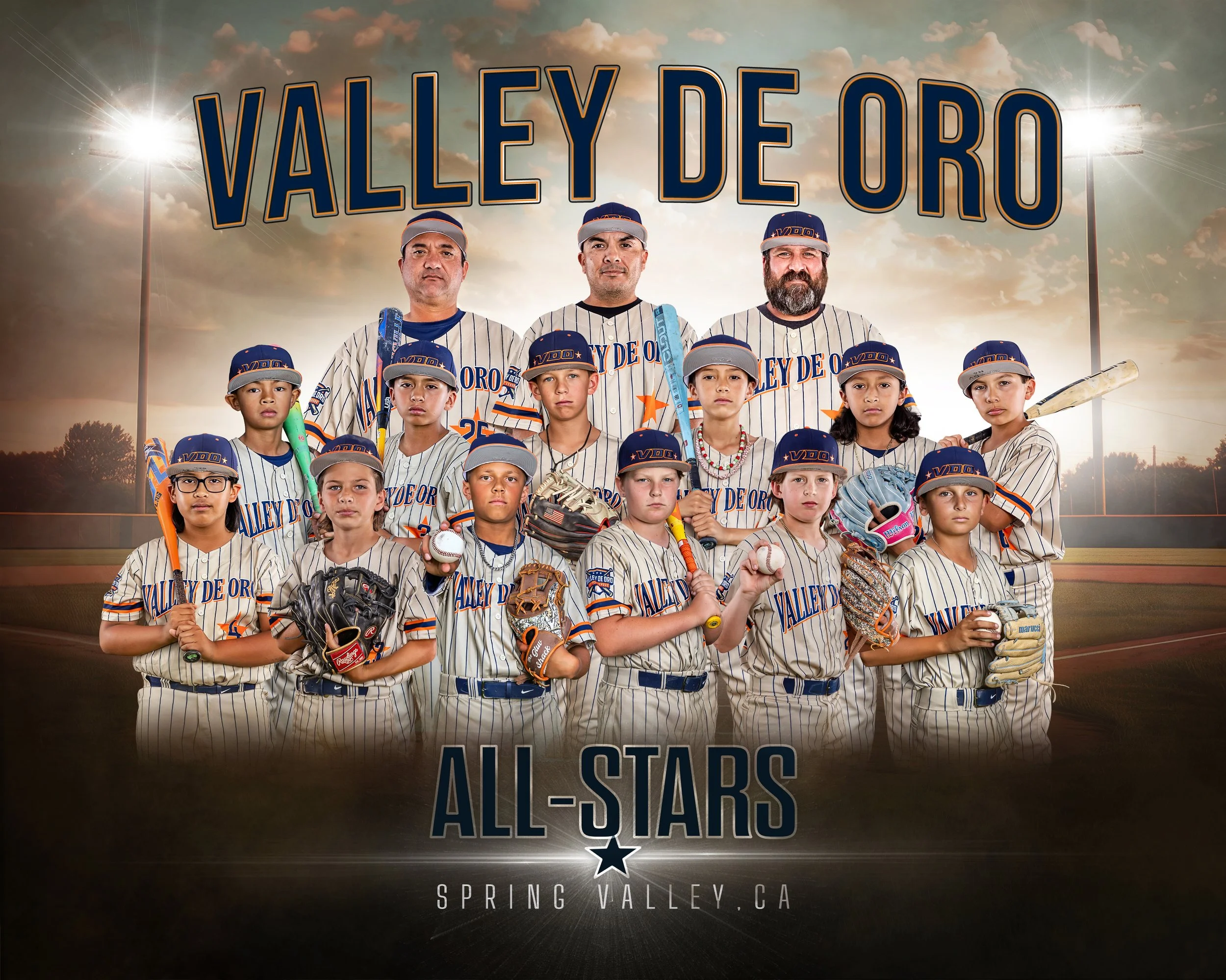 Group of children and adults in baseball uniforms posing on a baseball field under cloudy sky, with a large sign behind them reading 'Valley of De Oro All-Stars' and stadium lights in the background.