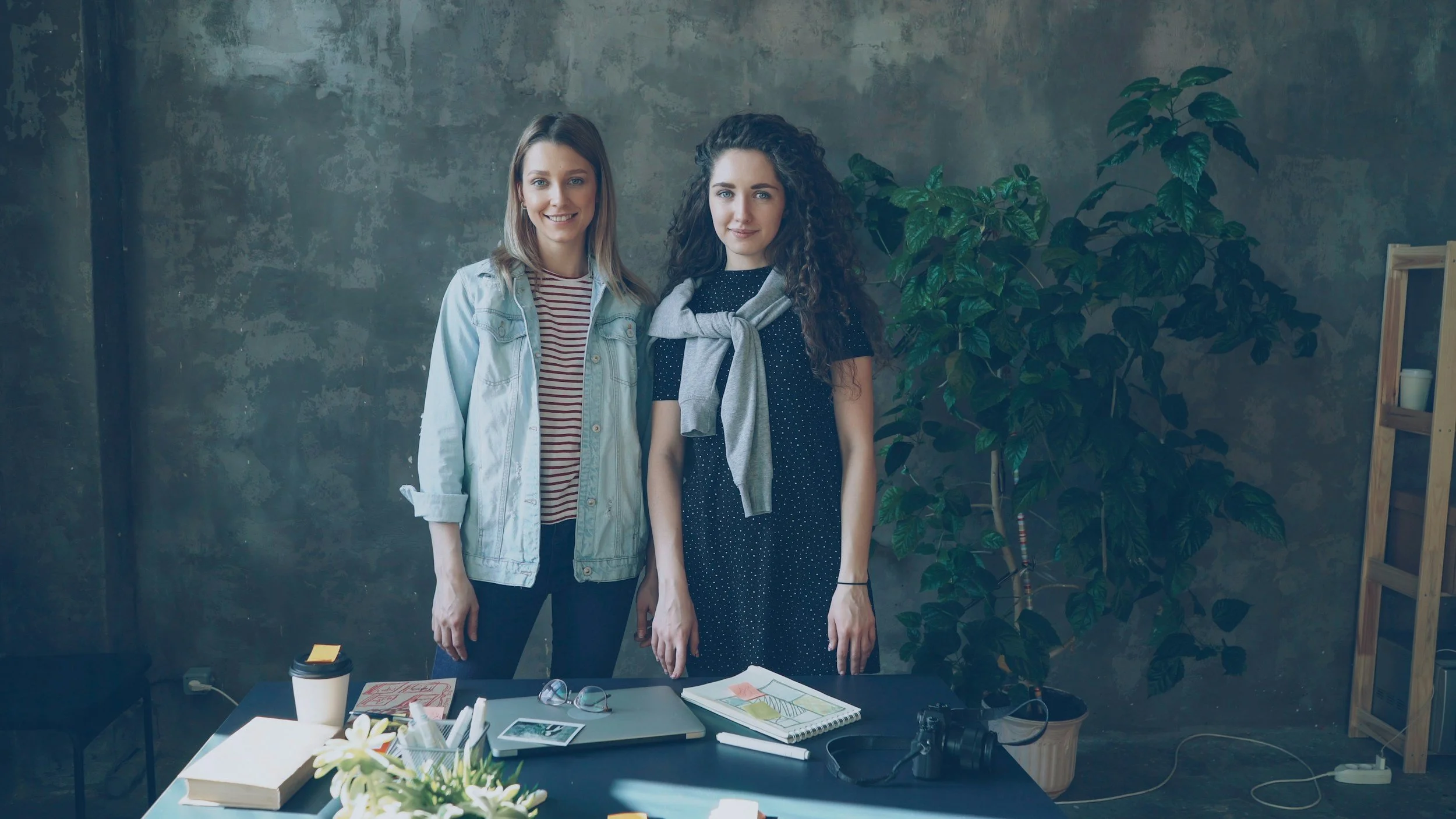 Two women standing behind a table in an indoor setting with a concrete wall and large leafy plant in the background. The table has various papers, books, a coffee cup, and a camera.