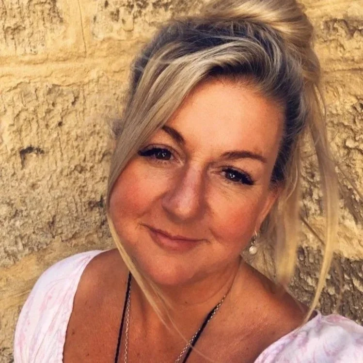 A woman with blonde hair styled in loose waves, smiling, with a beige textured stone wall background.