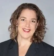 A woman with curly brown hair wearing a dark blazer and red lipstick, smiling against a light gray background.