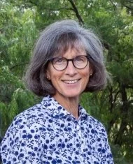 A woman with gray hair and glasses smiling outdoors, wearing a blue and white patterned blouse, with green foliage in the background.