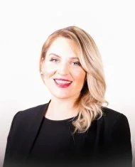 Headshot of a woman with blonde hair, wearing a black blazer, smiling, against a white background.
