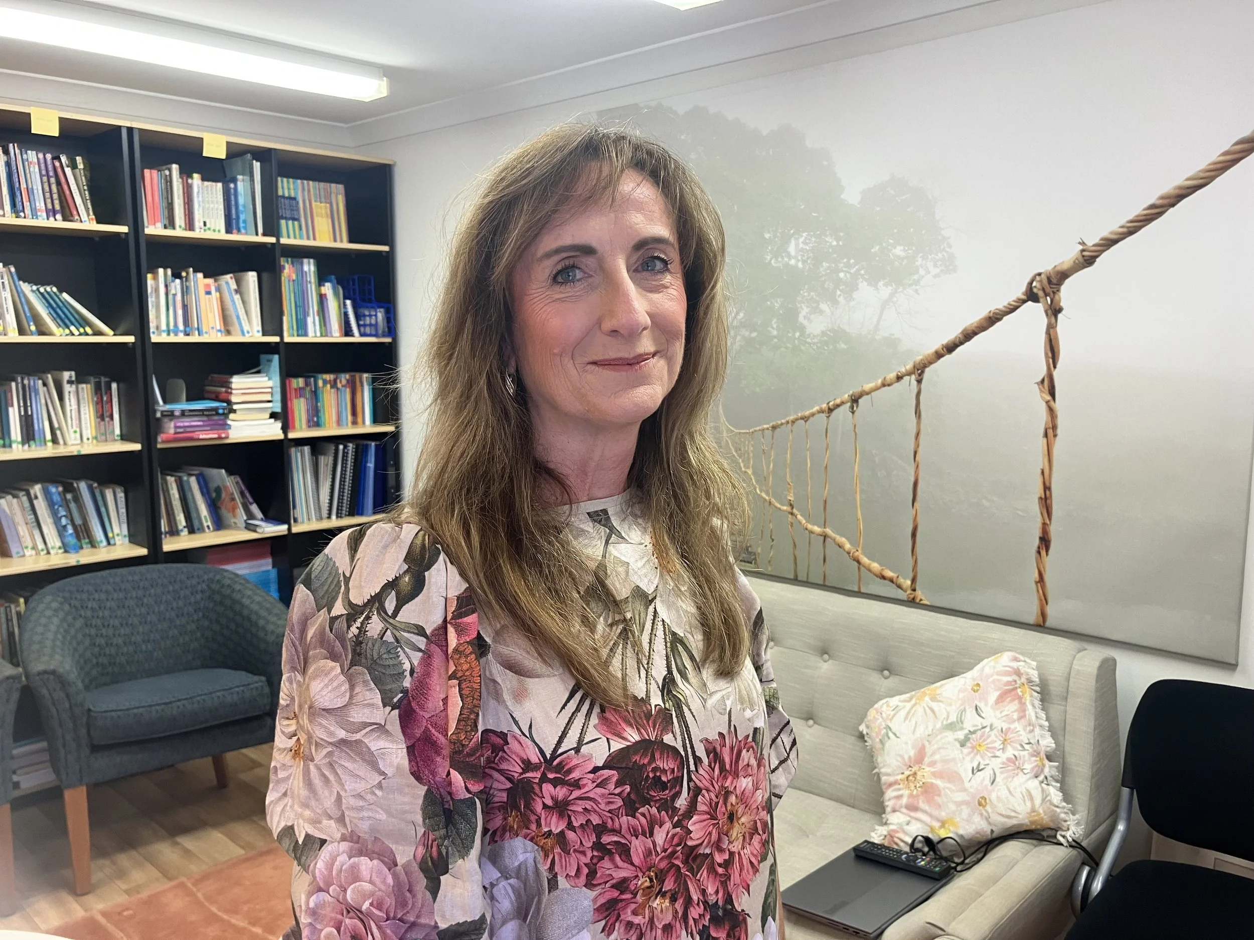 A smiling woman with long brown hair wearing a floral blouse standing in a room with a bookshelf filled with books, a blue armchair, and a wall with a foggy forest scene mural.