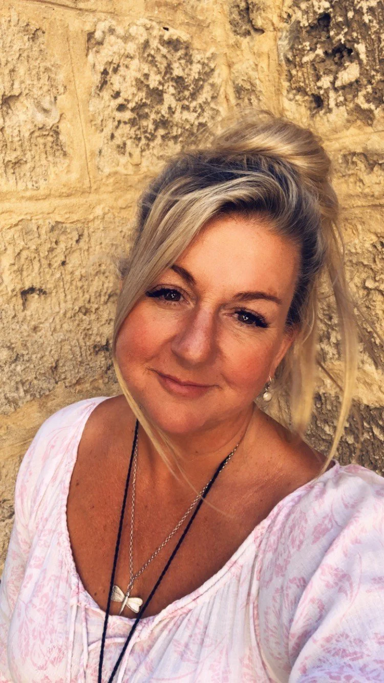 A woman with blonde hair styled in a high bun and loose strands, smiling, against a textured stone wall. She is wearing a light-colored top with a subtle pink pattern, layered necklaces, and earrings.