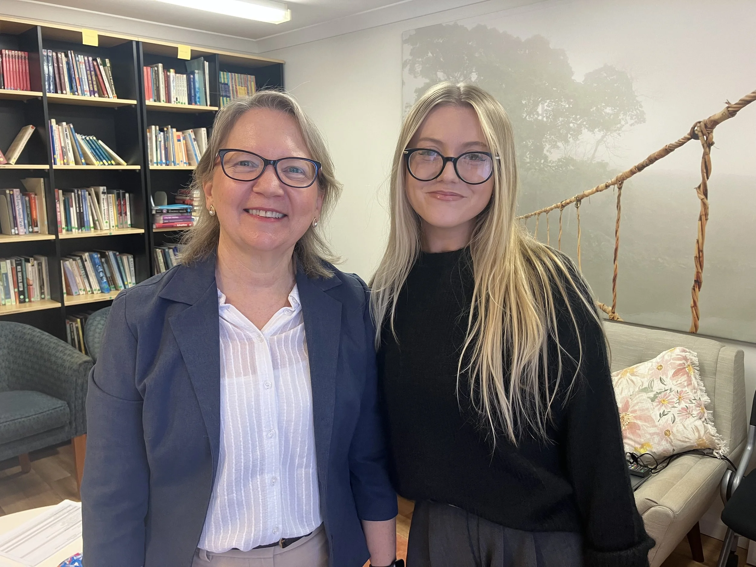 Two women with glasses smiling and standing close together in an indoor setting with a bookshelf and a wall mural of trees in the background.