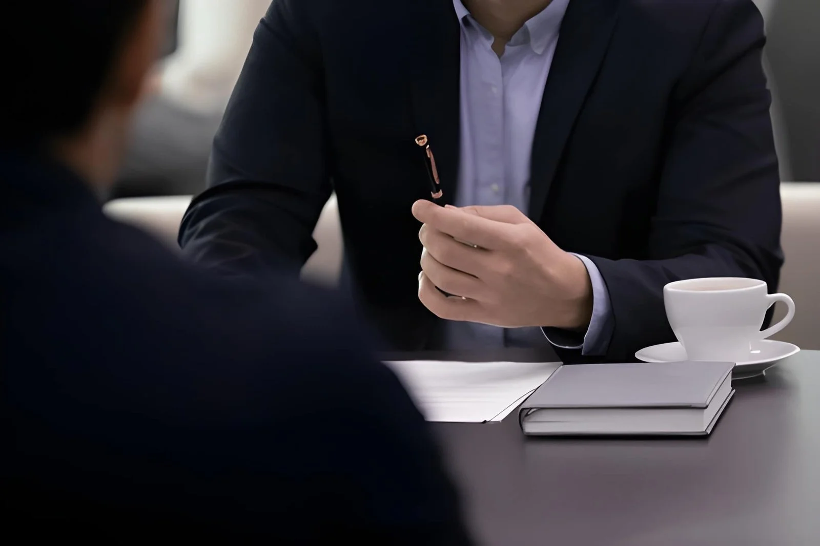 A person in a dark suit sitting at a table with a white cup and saucer, a closed notebook, and a notepad, engaged in conversation with another individual; the person is holding a pen.