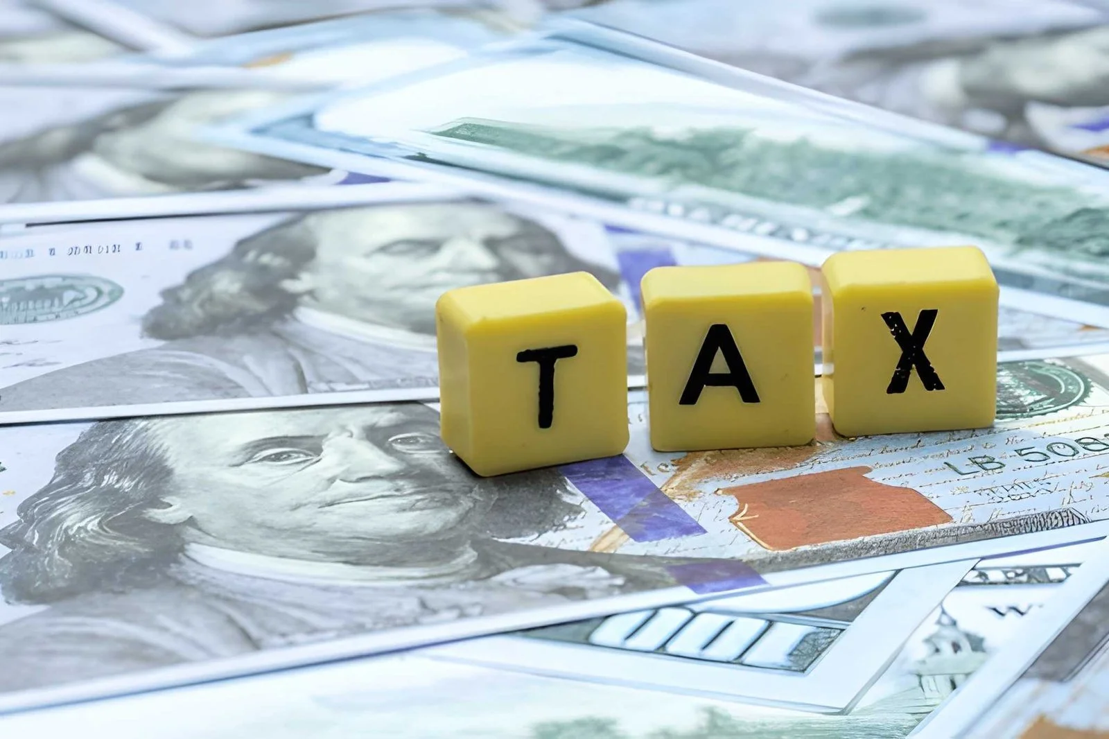 A pile of US $100 bills with yellow blocks spelling out 'TAX' placed on top.