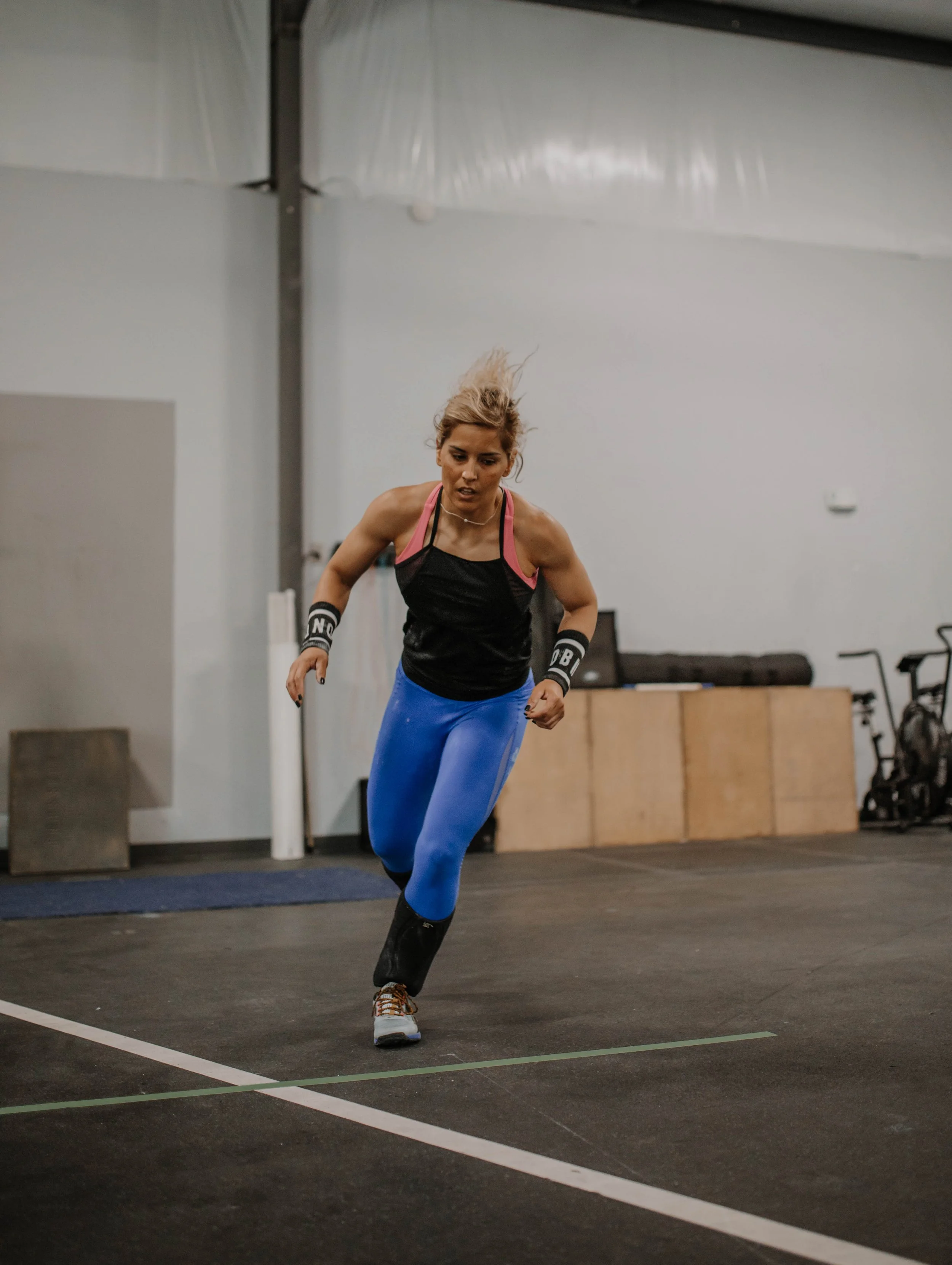 A woman running in an indoor gym, wearing blue leggings, black top, and wristbands.