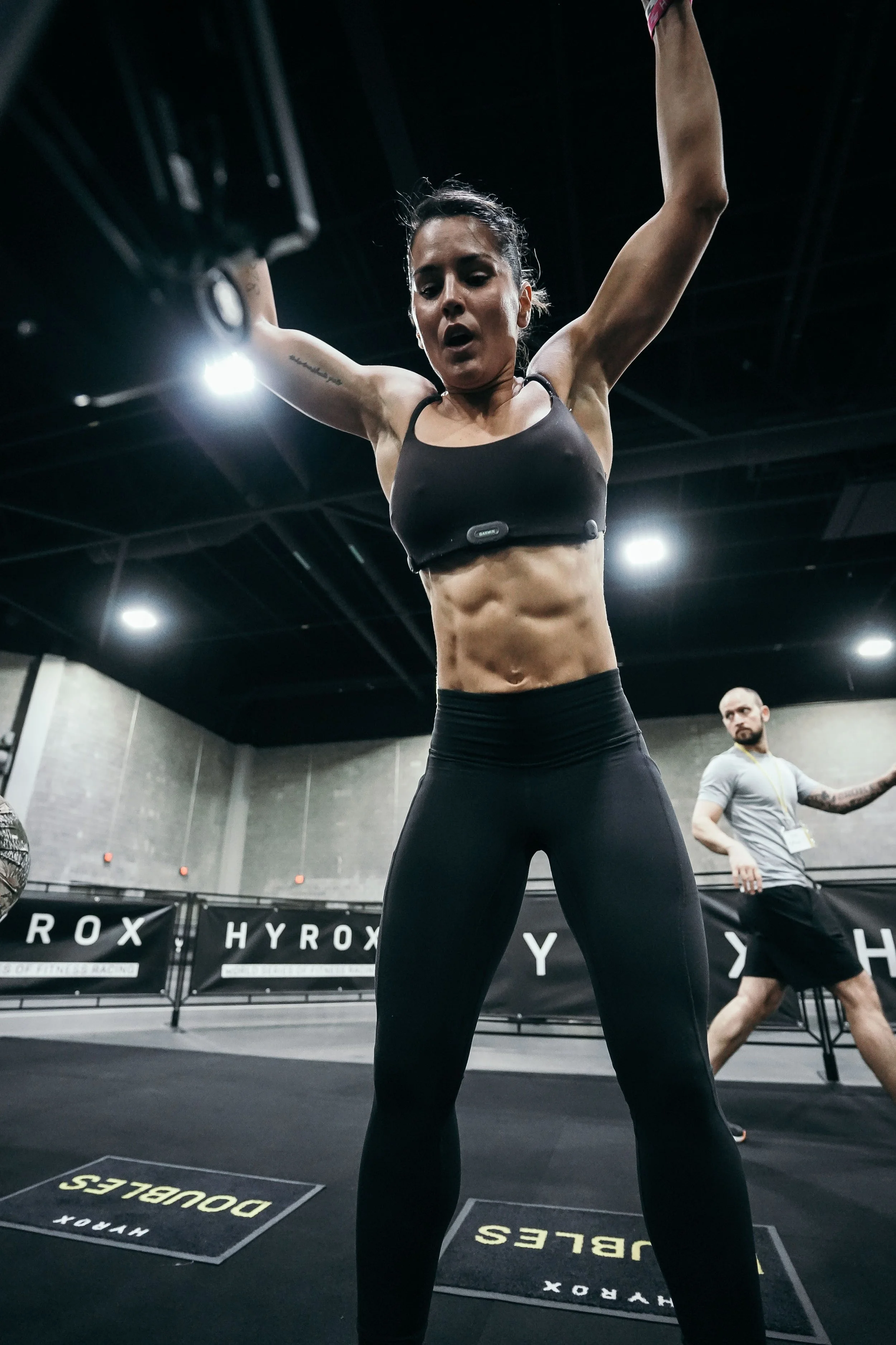 A woman in athletic clothing doing an individual Hyrox race, with a man in the background.