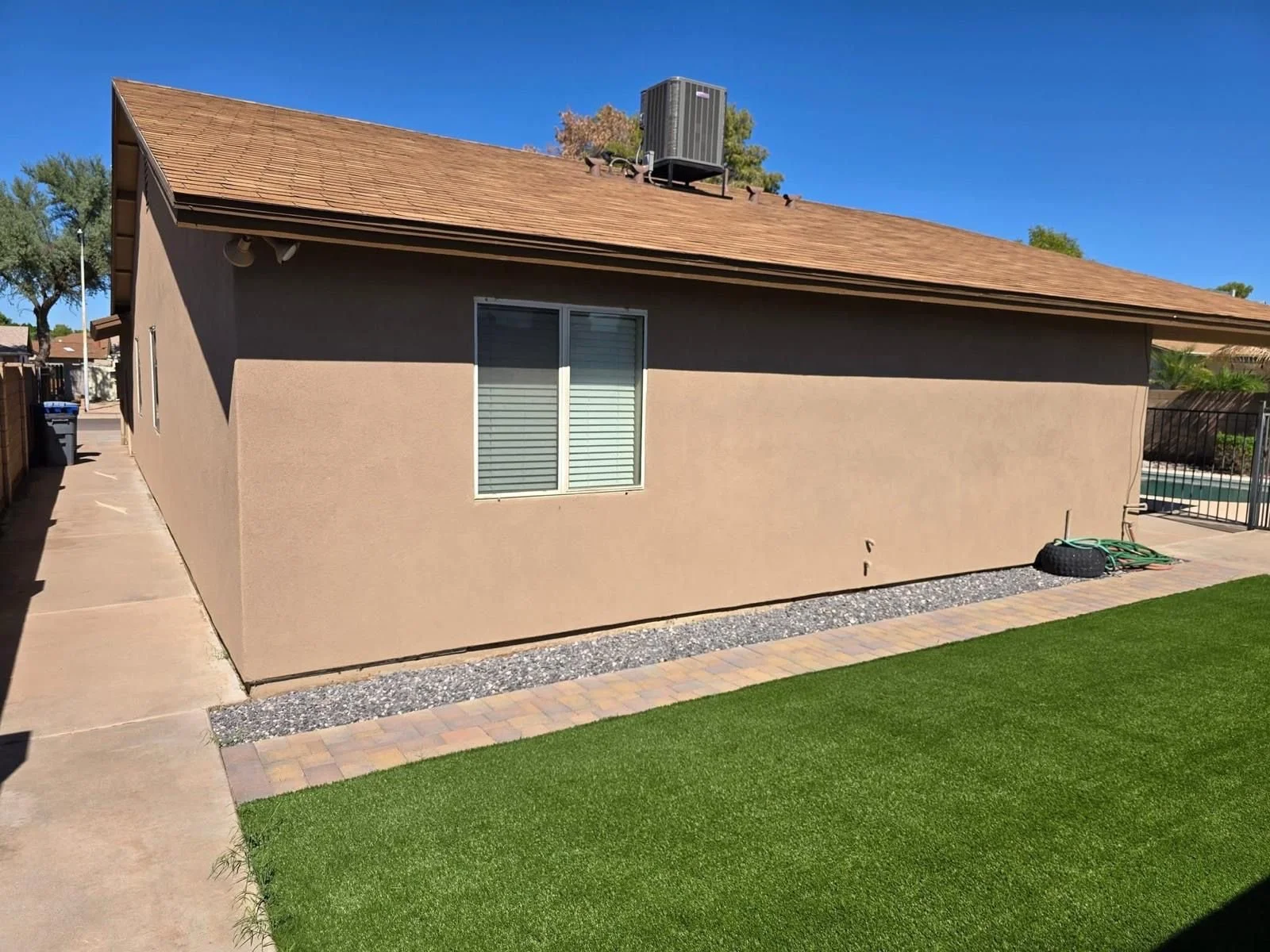 Backyard of a house with a beige stucco wall, a window with blinds, a concrete walkway, and a green artificial turf lawn. There is a tire and garden hose on the ground near the wall, and a chain-link fence on the right.