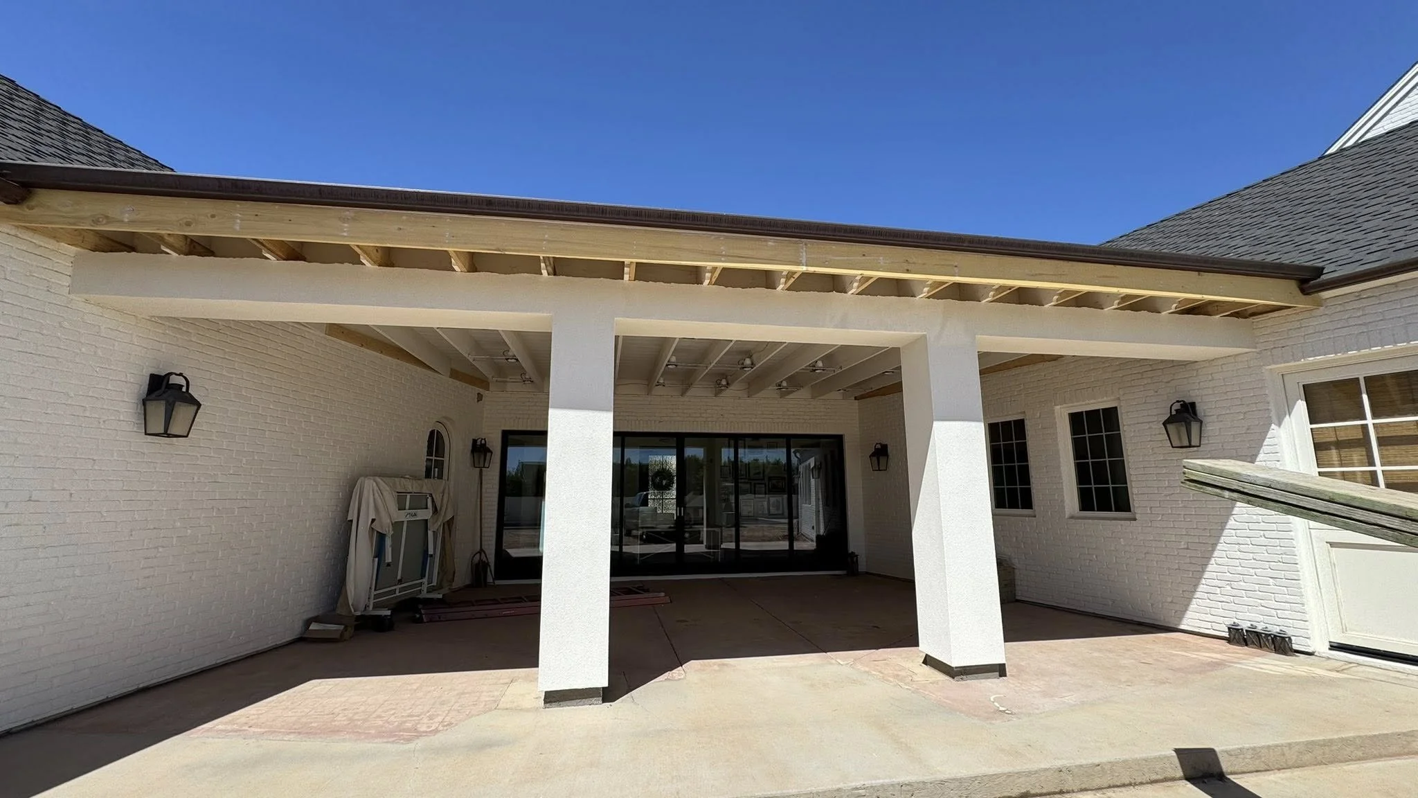 Back patio area with white brick walls, sliding glass door, and exterior wall lantern lights, under construction with exposed roof framing and no finished ceiling.