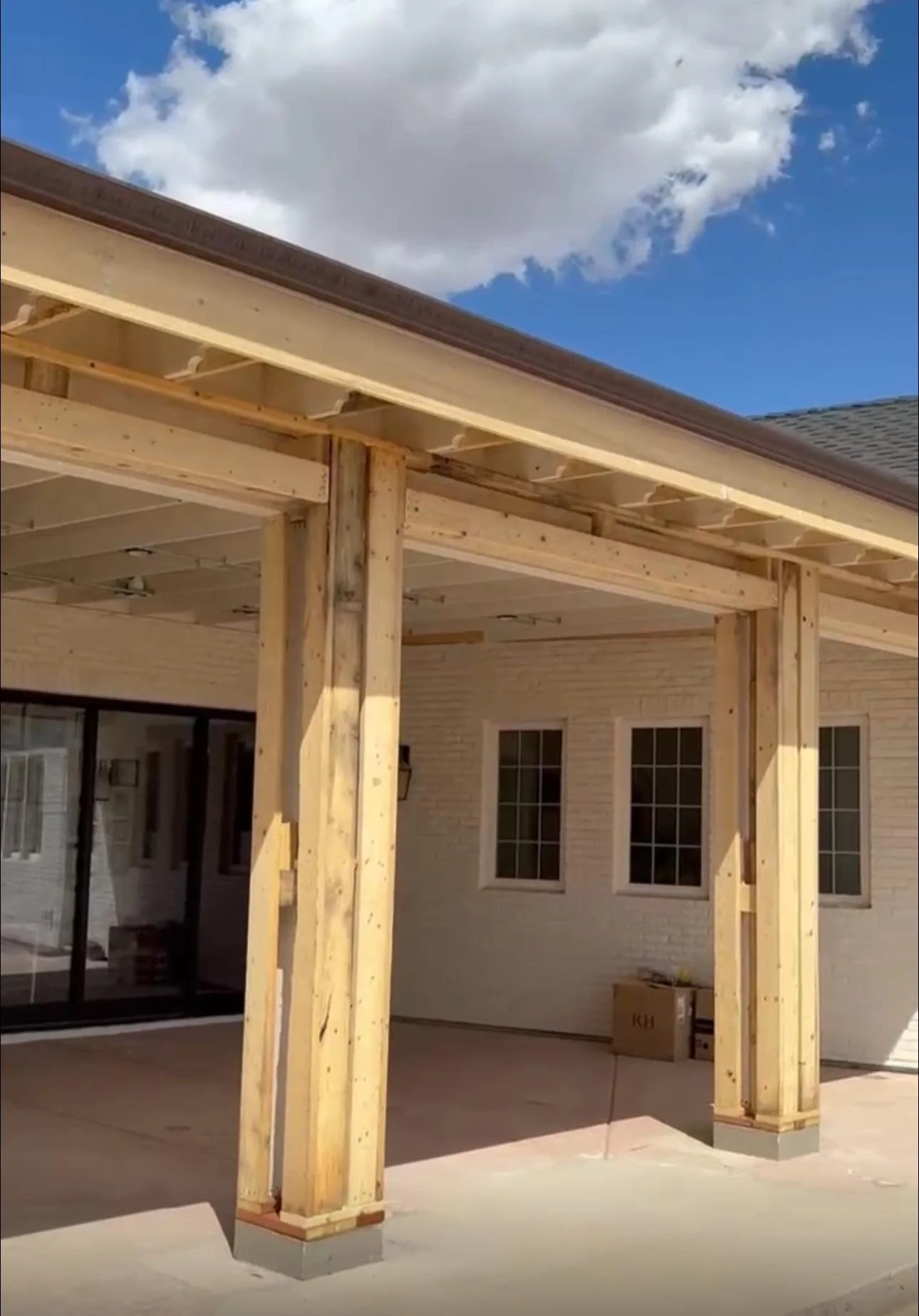 Unfinished porch construction with wooden beams and posts, part of a house with white brick exterior, several windows, and a cloudy blue sky above.
