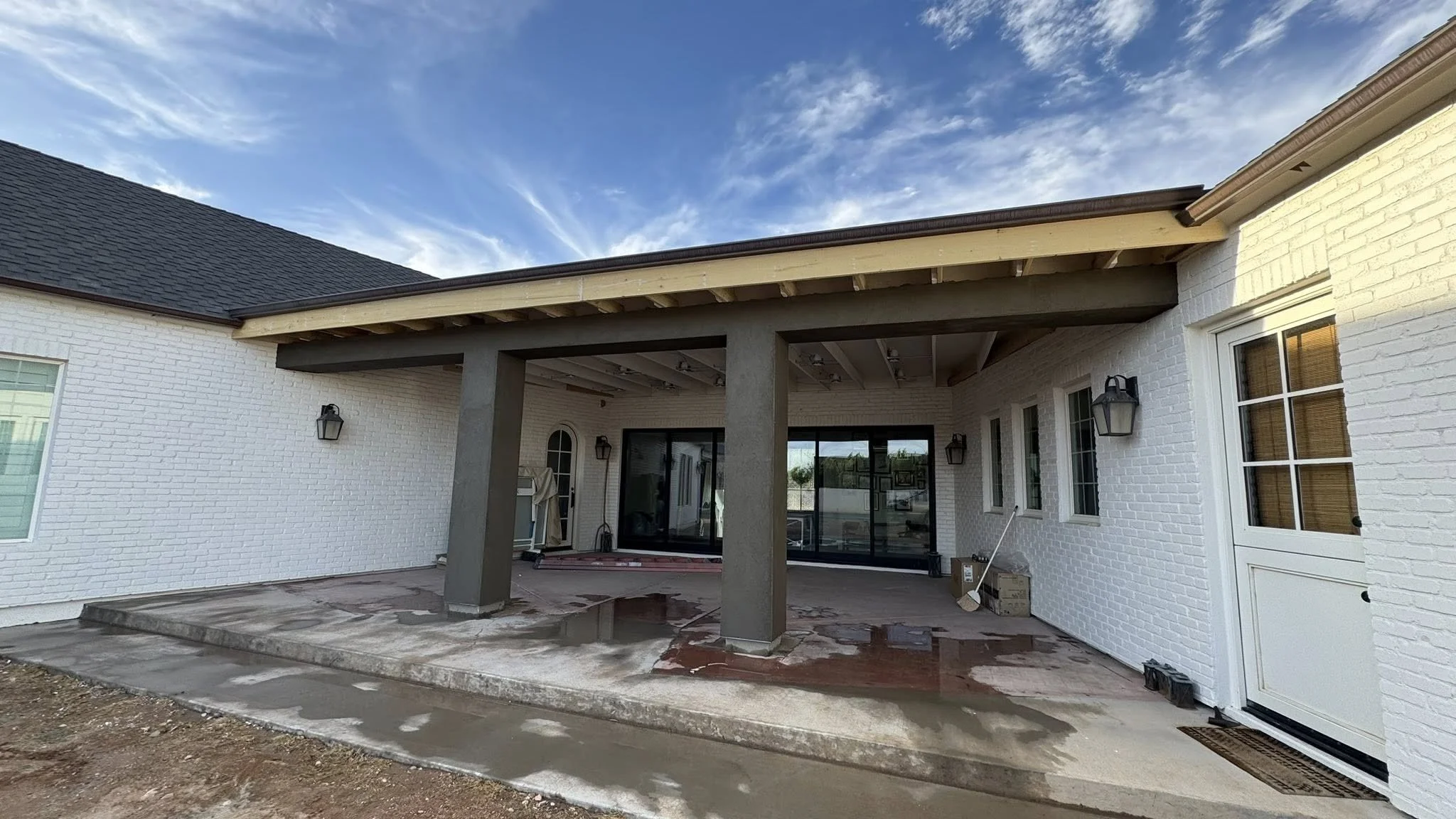 Back porch area of a house under construction with a concrete floor, white brick walls, black sliding glass doors, and exterior wall-mounted lanterns. Construction materials and tools are visible, and the sky is partly cloudy.