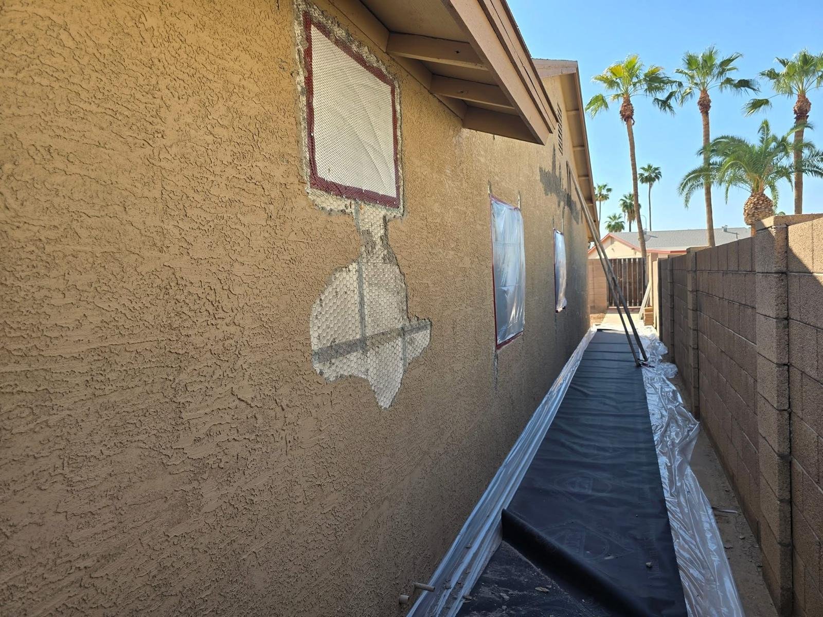 Side view of a house wall undergoing repair, with a section of stucco removed, exposing mesh underneath. Windows covered with plastic sheets, with a walkway covered in plastic sheeting leading to a backyard. Tall palm trees are visible in the background.