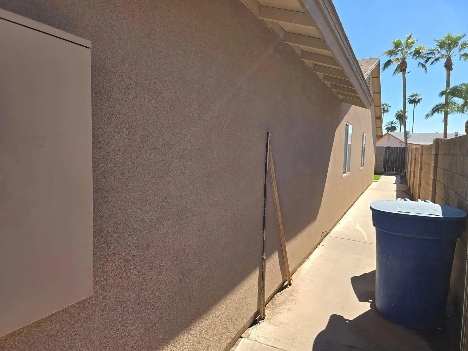 Side of a house under construction with a dirt wall, a blue trash bin, a wooden construction tool leaning against the house wall, and palm trees in the background on a sunny day.