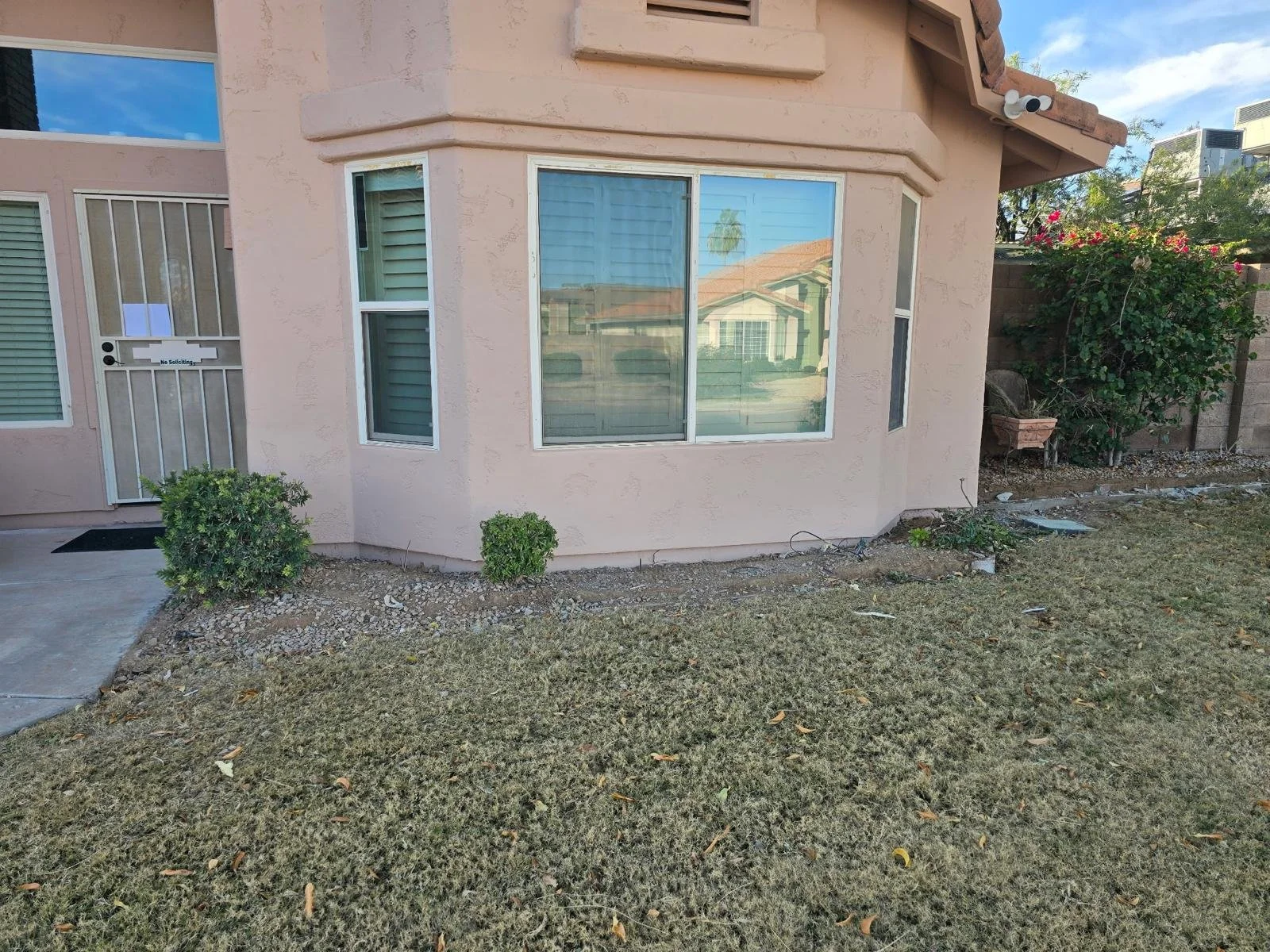 The exterior of a peach-colored house with three large windows, a side door with a 'No Soliciting' sign, small bushes, and a yard with patchy grass and scattered leaves.