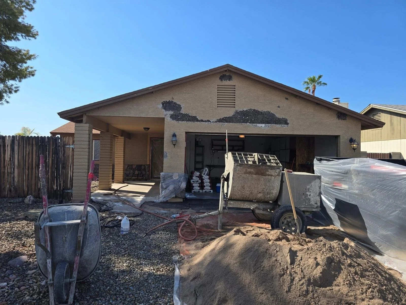 Front yard of a house under renovation with a pile of sand, construction equipment, and building supplies in the yard.