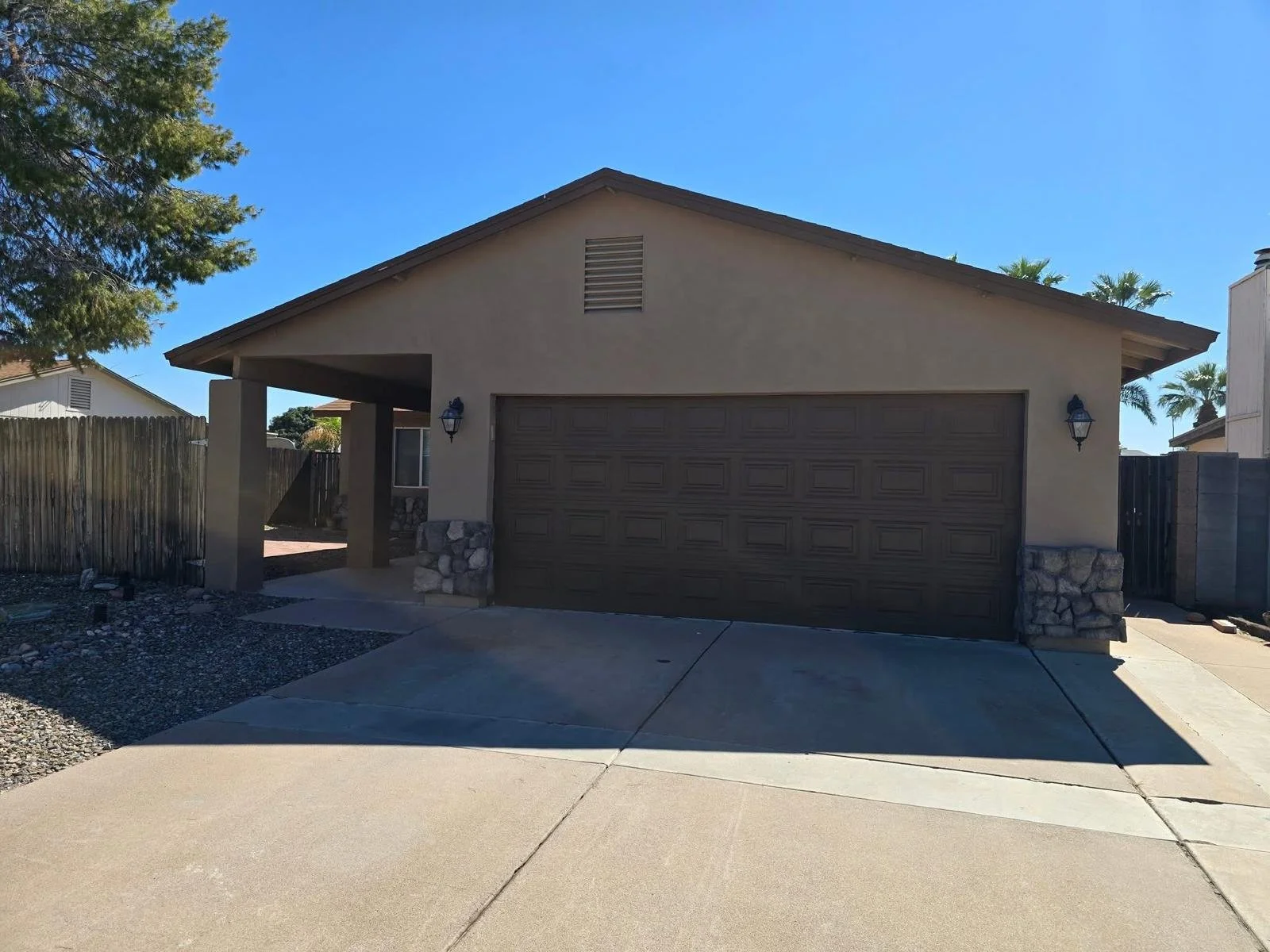 Front view of a house with a garage door, lit by two wall-mounted lanterns, surrounded by a driveway and a wooden fence, with trees and blue sky in the background.