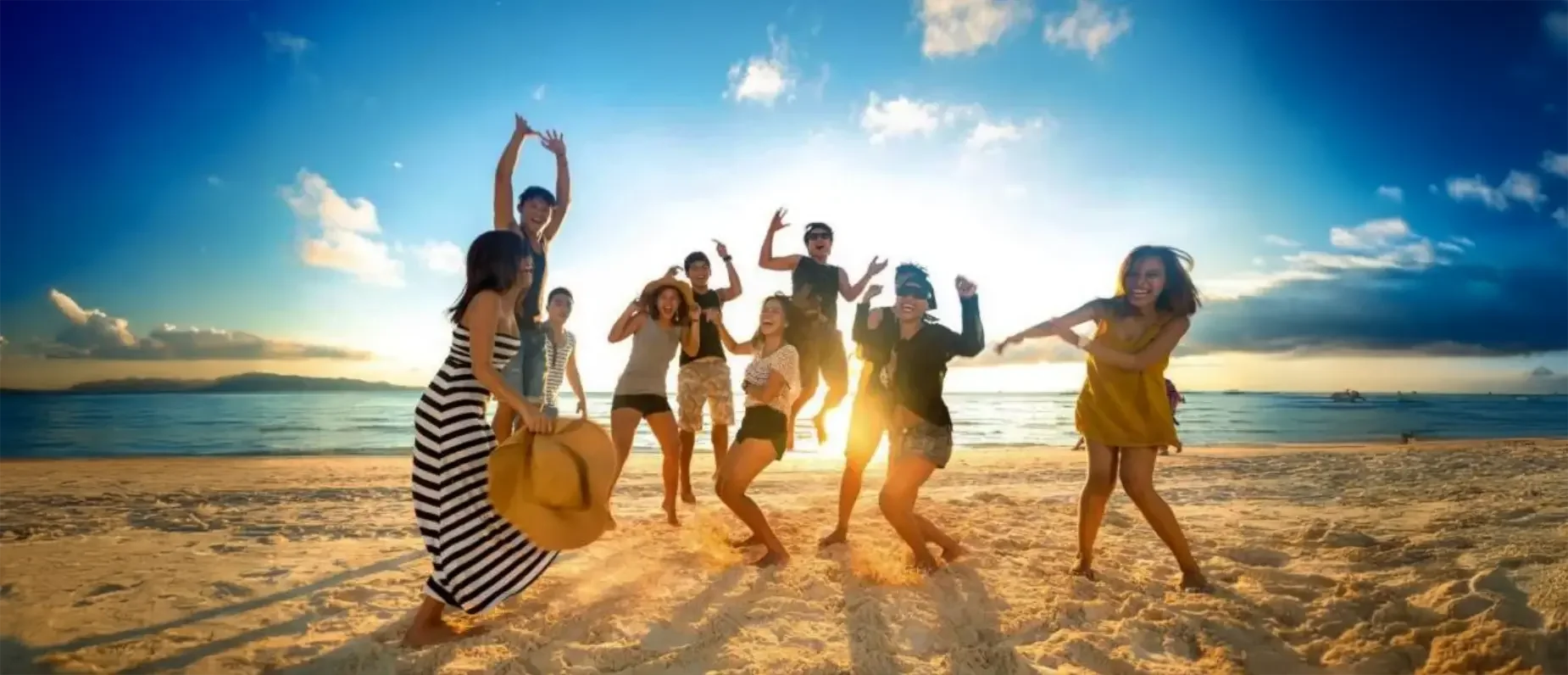 Group of friends having fun on the beach at sunset, jumping and smiling, with the ocean and sky in the background.