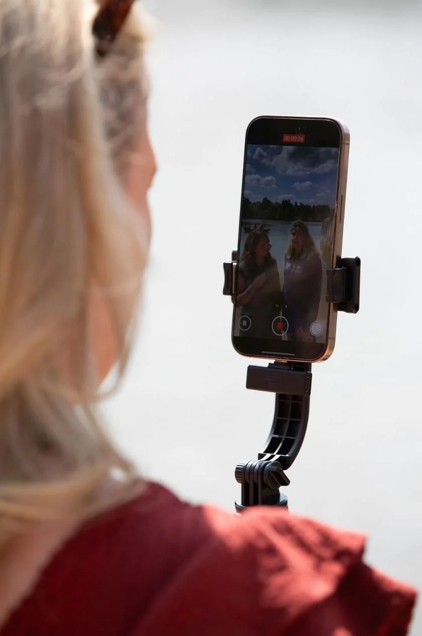 A woman with blonde hair in a red shirt records a video of two young women outdoors by a body of water, with a partly cloudy sky in the background.