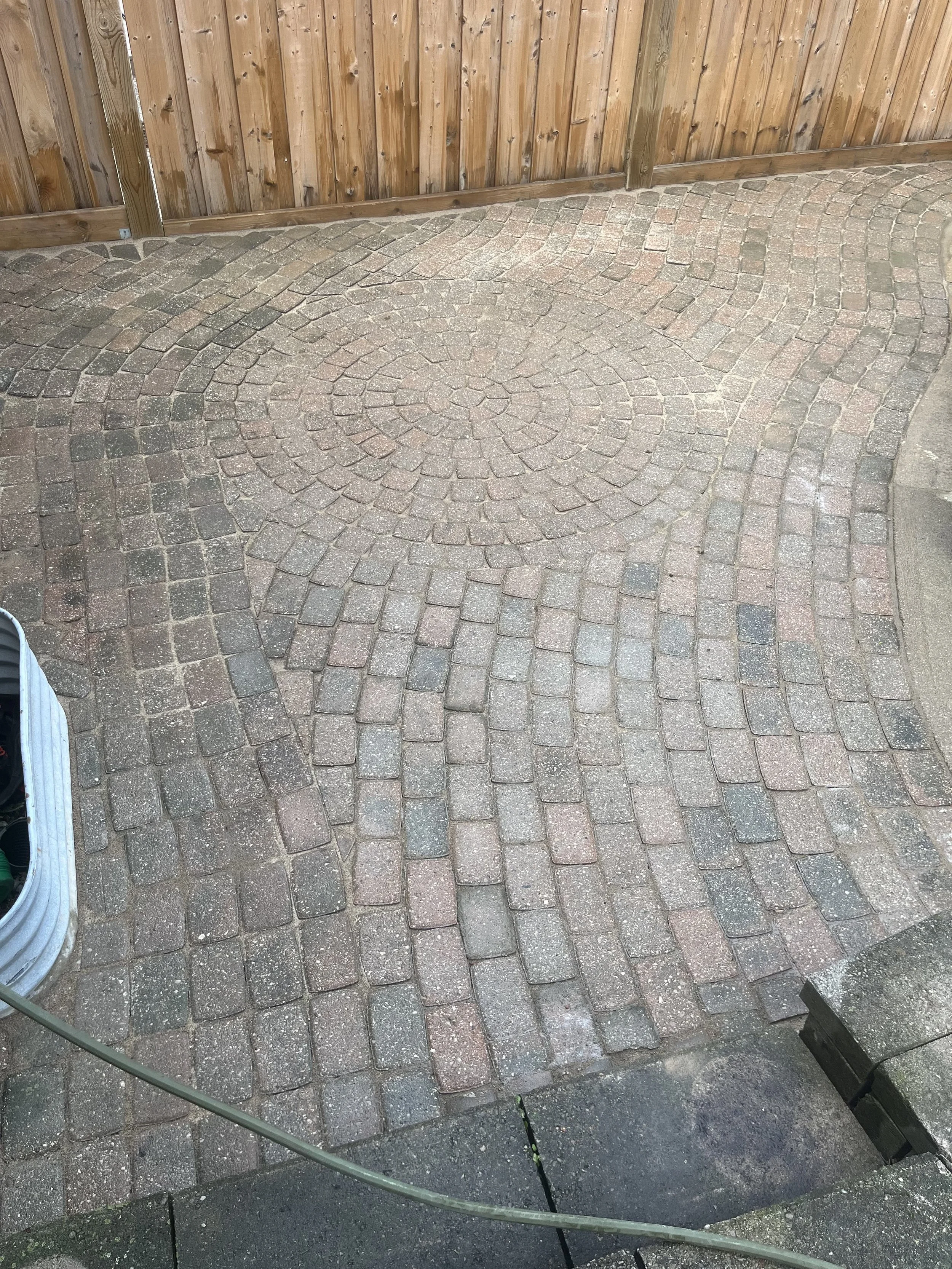 A brick patio with a circular pattern in the center, surrounded by a curved pattern. A wooden fence is in the background, and there are some gardening tools and a garden hose visible in the bottom left corner.