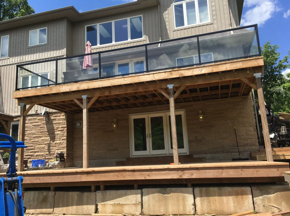 Unfinished wooden deck with support posts attached to a house with beige siding, located above a stone wall, under a partly cloudy sky.
