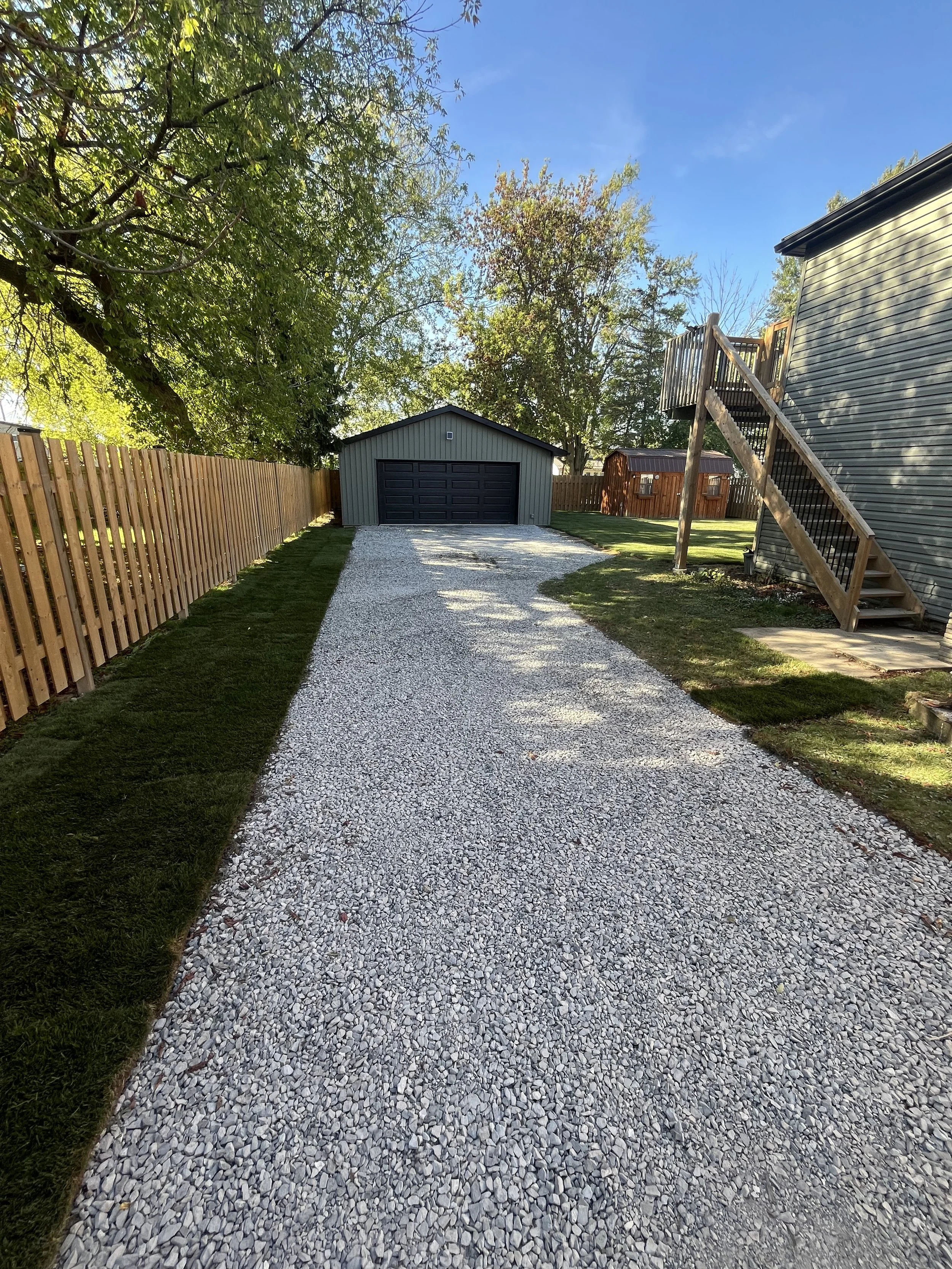 A gravel driveway leading to a dark green garage with black doors, bordered by wooden fences and a grassy lawn on both sides. There are trees overhead against a blue sky.
