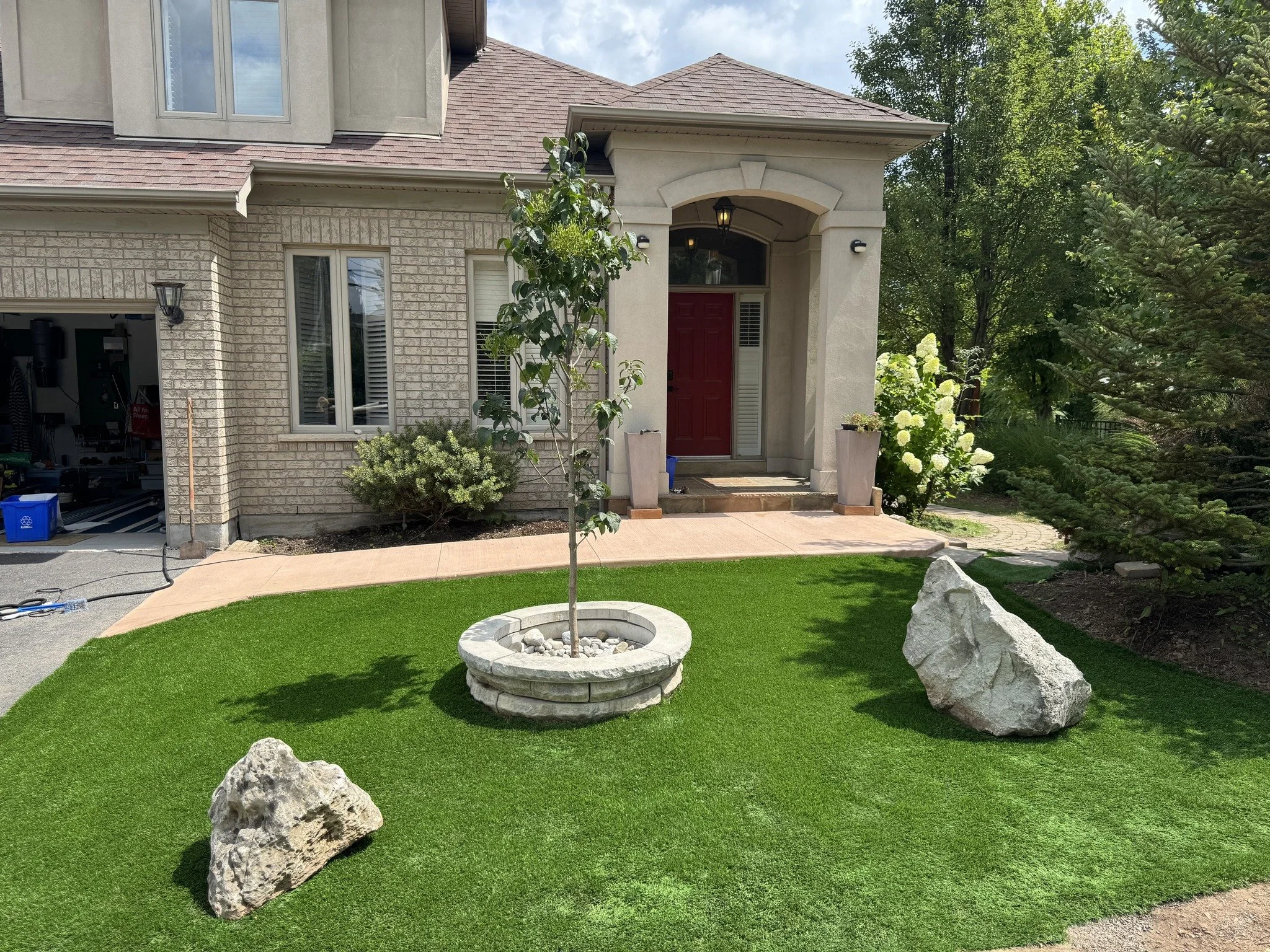 Front yard with artificial green grass, a young tree in a round stone planter, several large rocks, a porch with steps leading to a red door, a window with white shutters, and surrounding trees and shrubs.