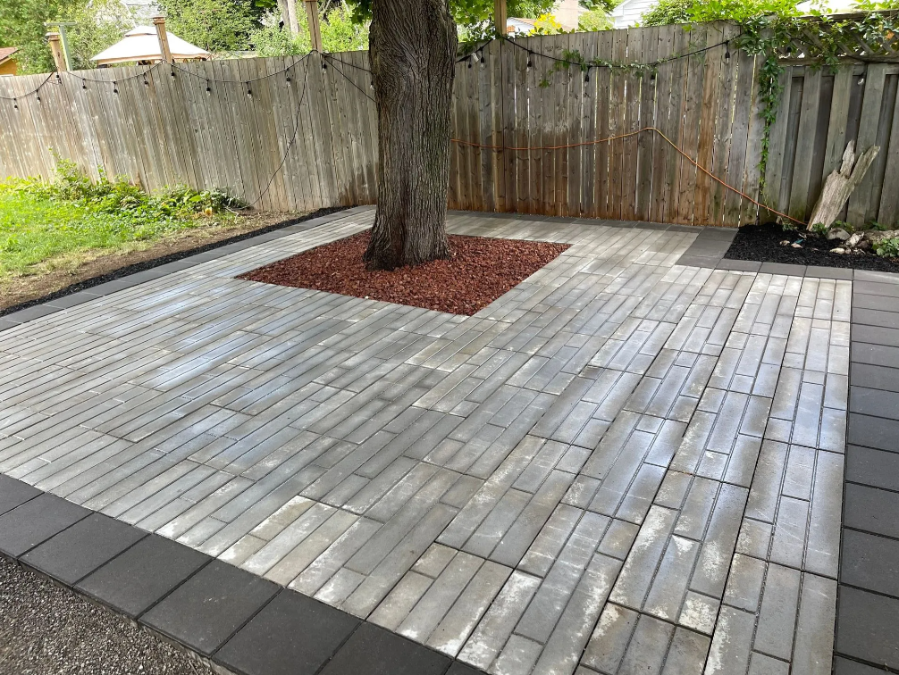 Patio with a large tree in the center, surrounded by pavers and bordered by black bricks, with a wooden fence and greenery in the background.
