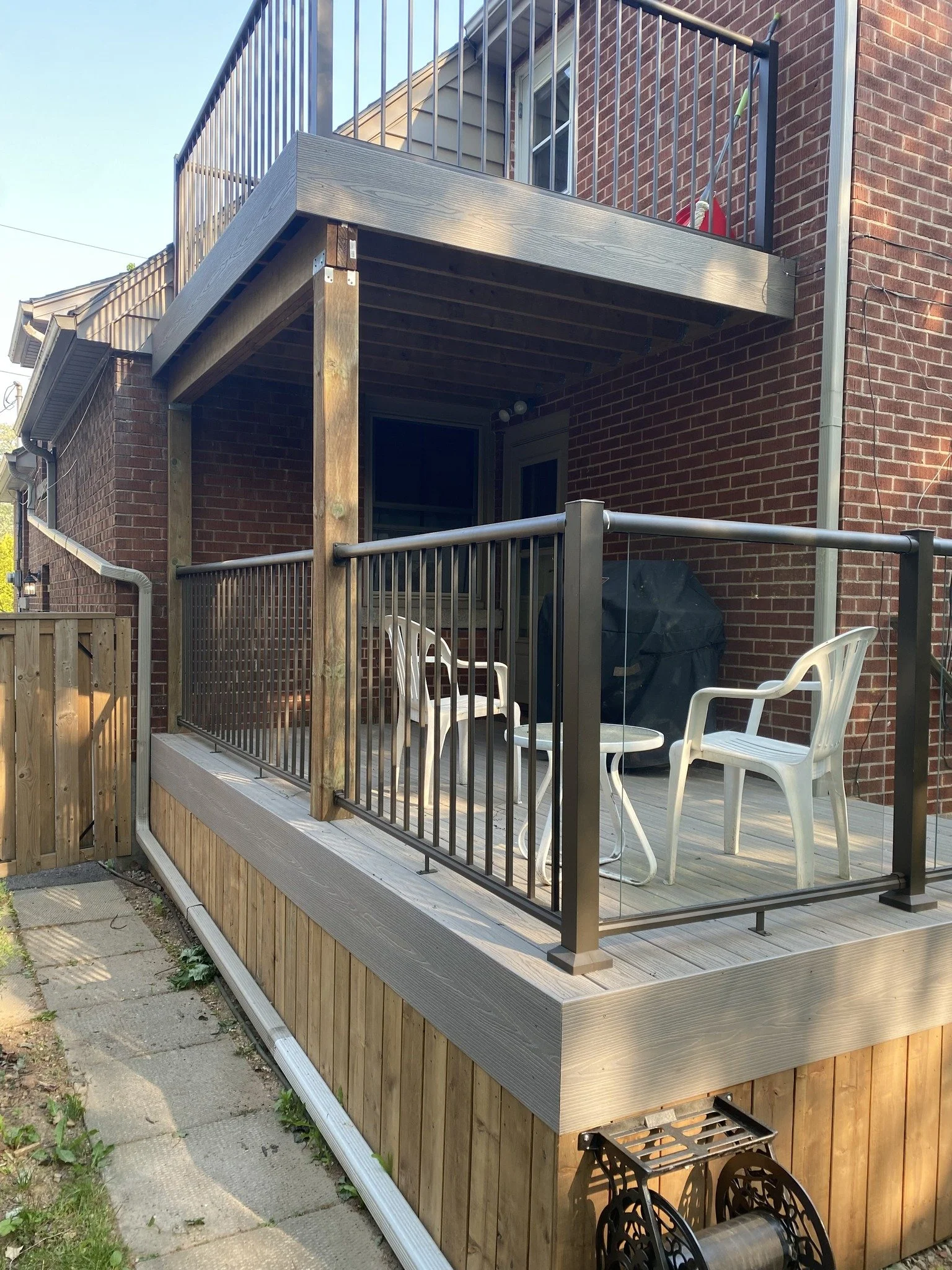 Backyard deck area with wooden flooring, protected by a black metal railing, attached to a brick house with a sliding door, outdoor chairs, covered grill, and cleaning tools visible.