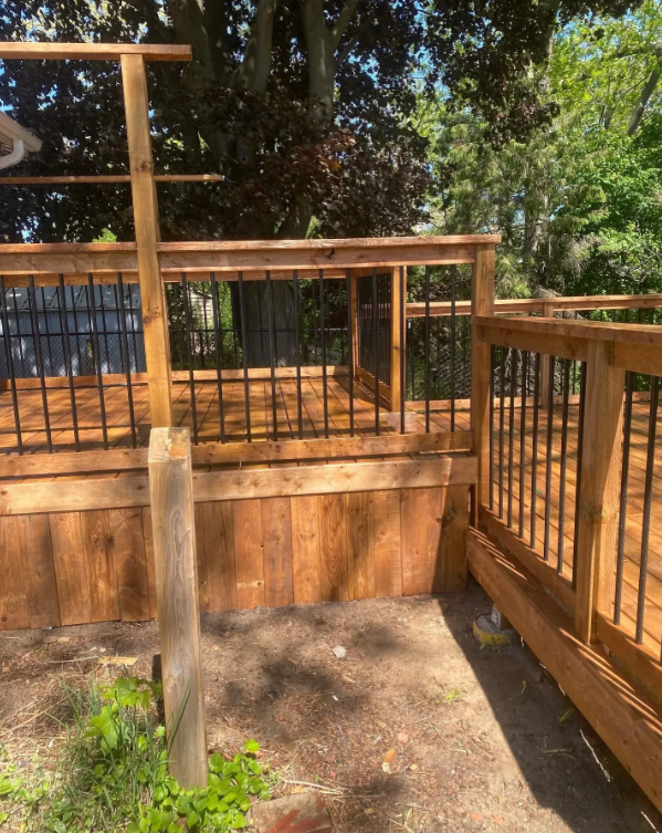 A newly built wooden deck with black metal railing, surrounded by trees and greenery, with sunlight creating shadows on the floor.