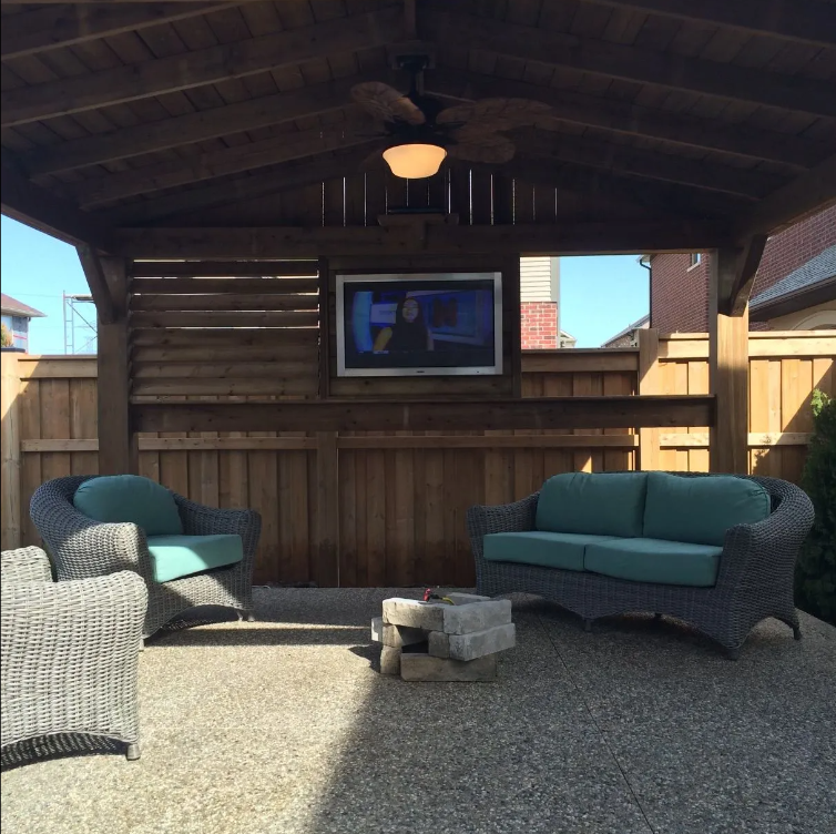 Outdoor covered patio with wicker furniture, including two armchairs and a loveseat with teal cushions. A small rectangular table made of concrete blocks is in the center. There is a flat-screen TV mounted on a wooden fence behind the furniture. The 