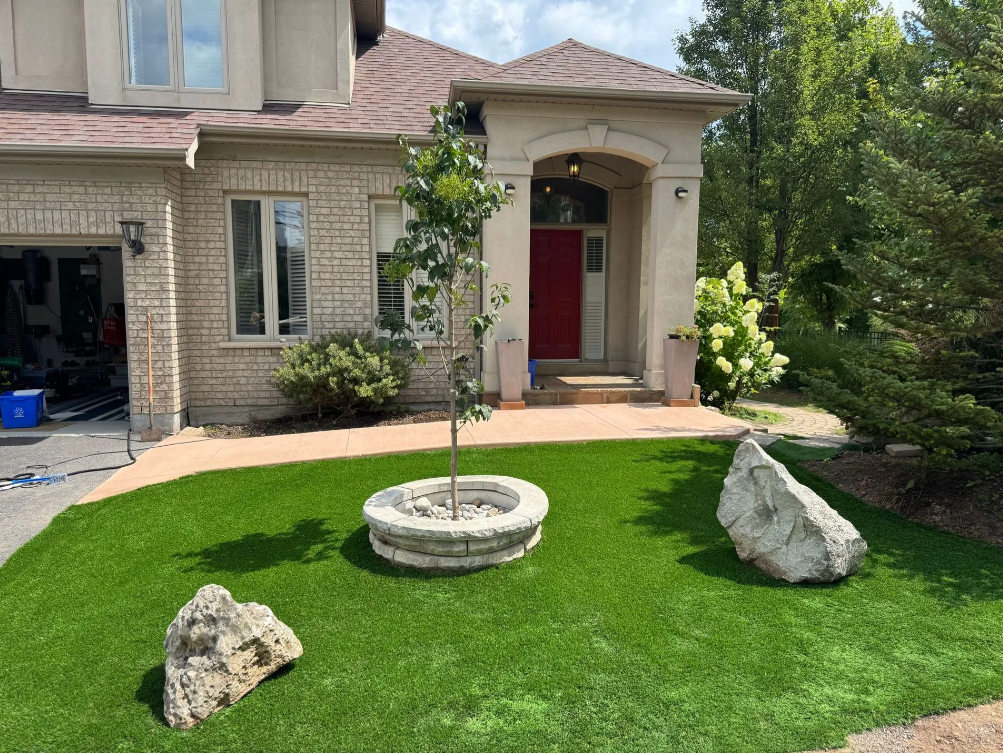 Front yard of a house with a small tree in a circular stone planter, three large rocks on a well-maintained grassy area, and a walkway leading to a red front door with white flowers nearby.