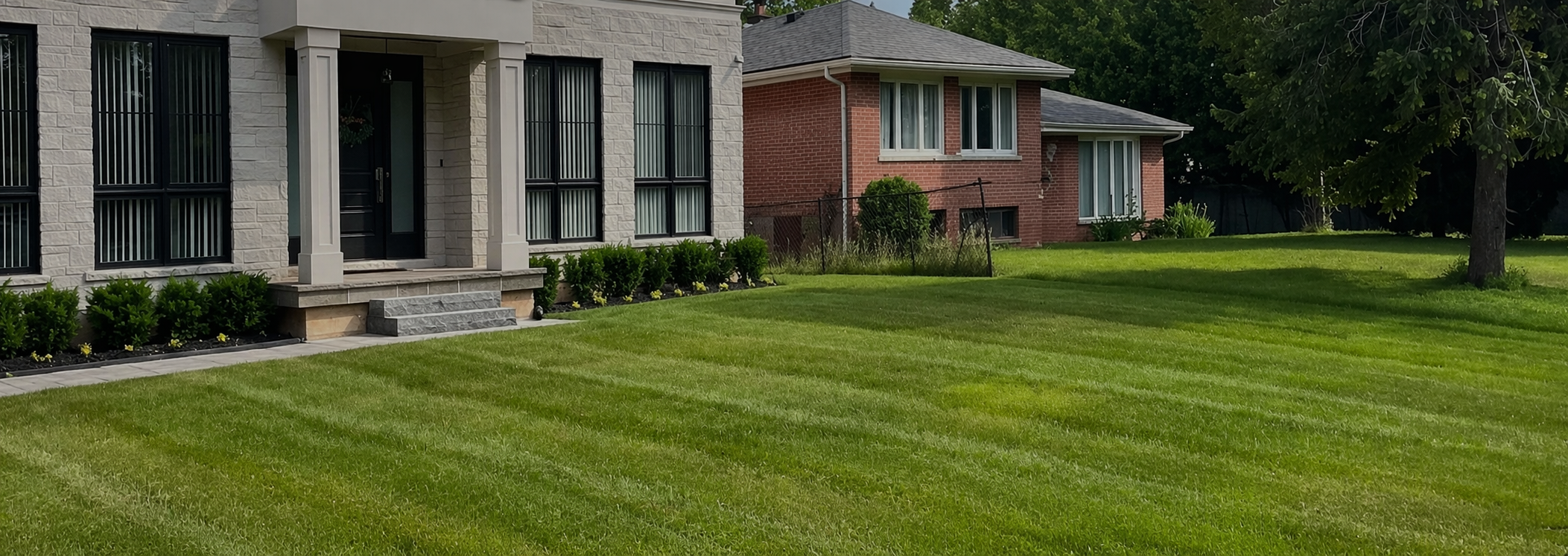 Front yard of a house with a well-maintained green lawn, an exterior of a house with white brick and large black window frames, steps leading to a black door, and a neighboring red brick house with large windows in the background. A tree and some bushes are also visible.