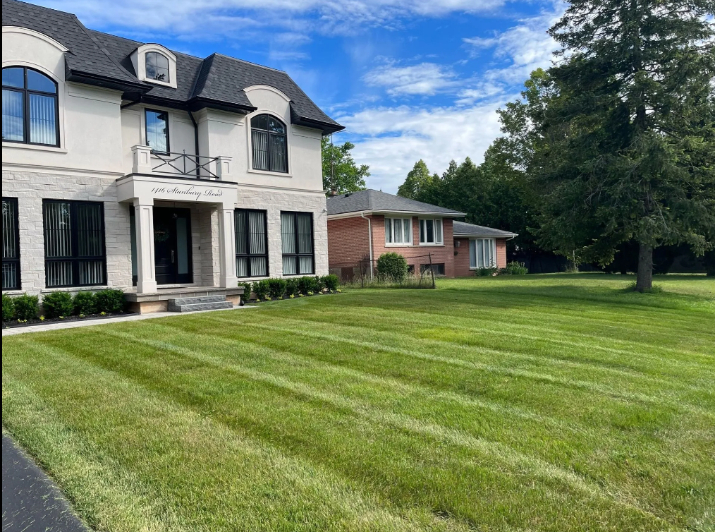 A large white two-story house with black roof and windows, a small balcony, and the address 1116 Stambury Road on the front, surrounded by a well-maintained lawn and a tree on the right, with a partly cloudy sky overhead.
