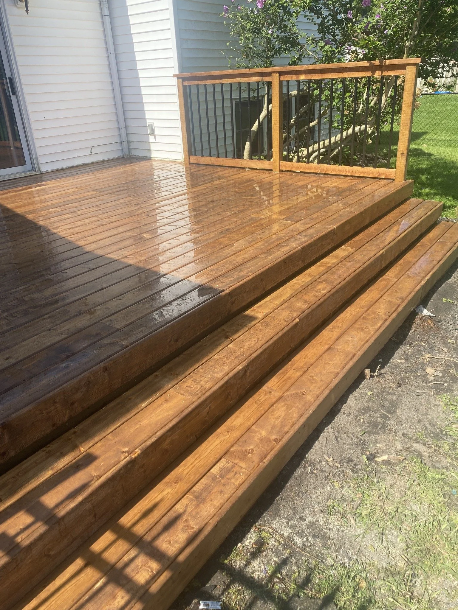 A freshly stained wooden deck with steps, adjacent to a white house with vinyl siding. The deck includes a railing made of wooden posts and black metal balusters, and is situated in a backyard with green grass, a tree with pink flowers, and a wire fe