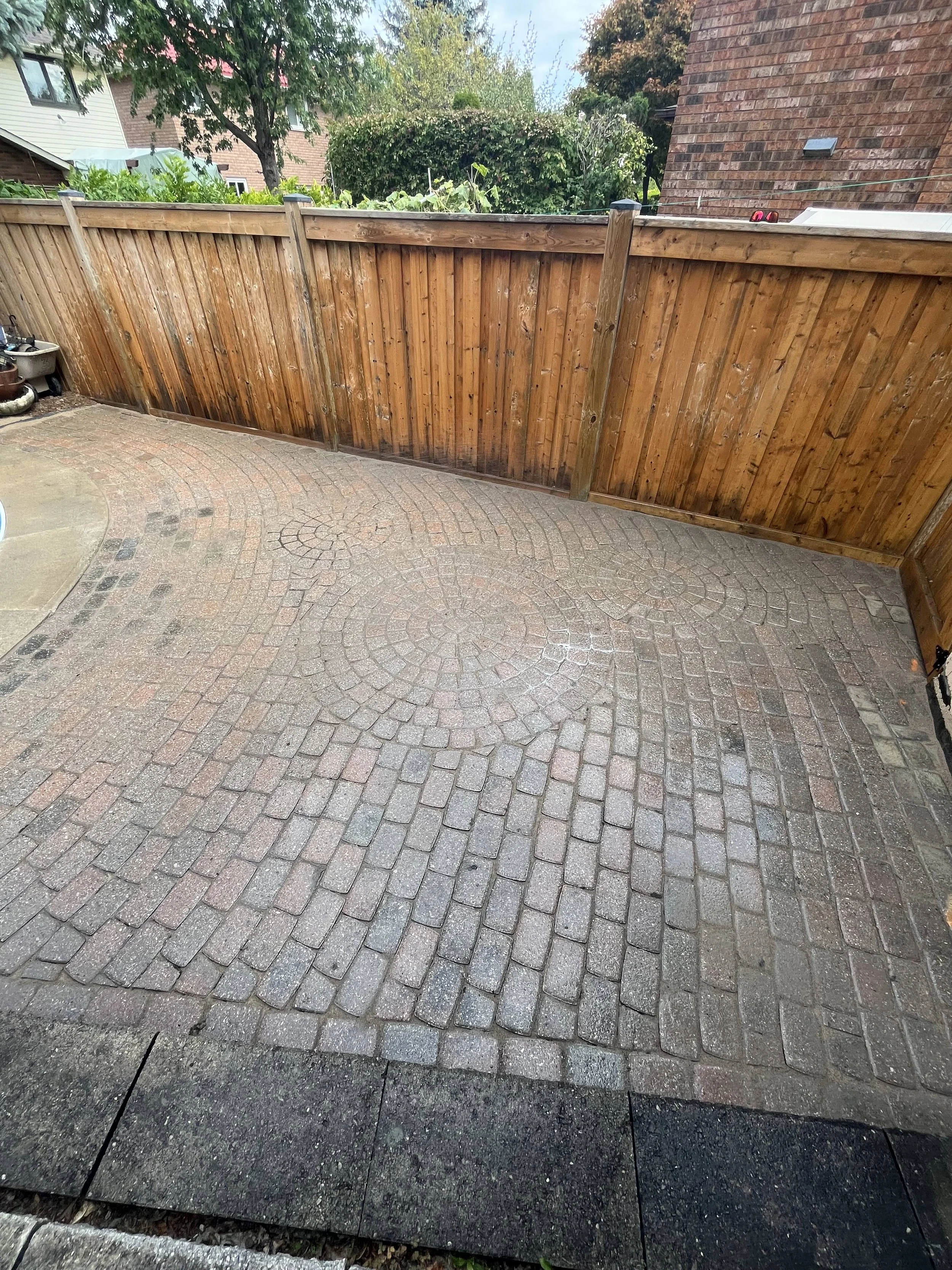 View of a backyard with a wooden fence, paved patio, and neighboring houses and trees.