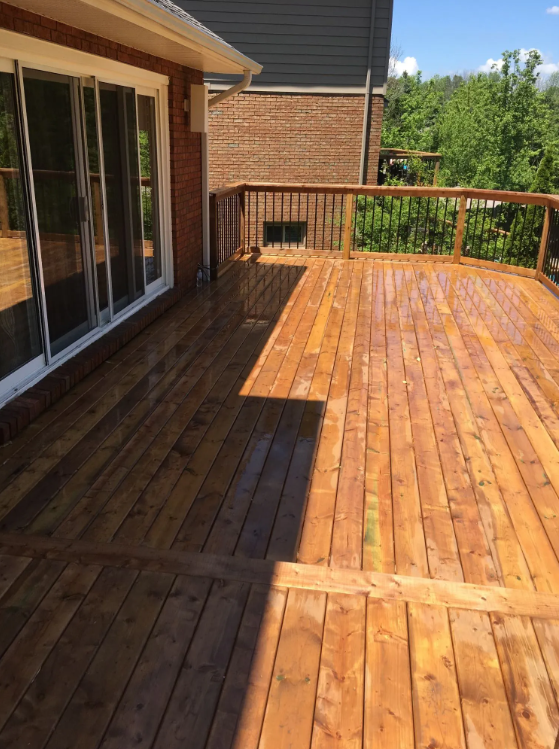 Freshly cleaned wooden deck attached to a brick house, with glass sliding doors leading inside, surrounded by green trees and a blue sky.