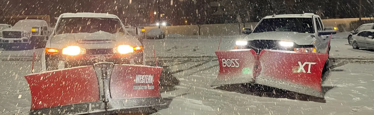 Two pickup trucks equipped with snow plows clearing snow in a parking lot during snowfall at night.