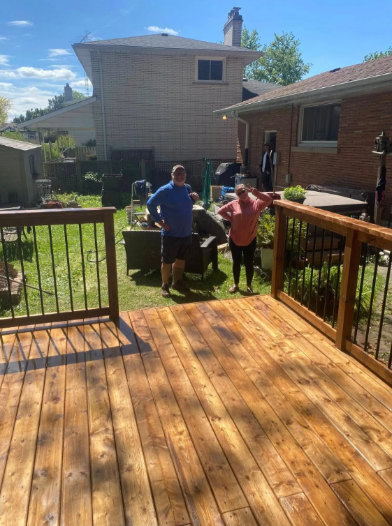 View of a backyard deck and patio area with two people standing. The deck has a wooden surface and black railing. Behind, there is a grassy area with outdoor furniture, gardening tools, and a barbecue grill. Two houses are visible in the background u